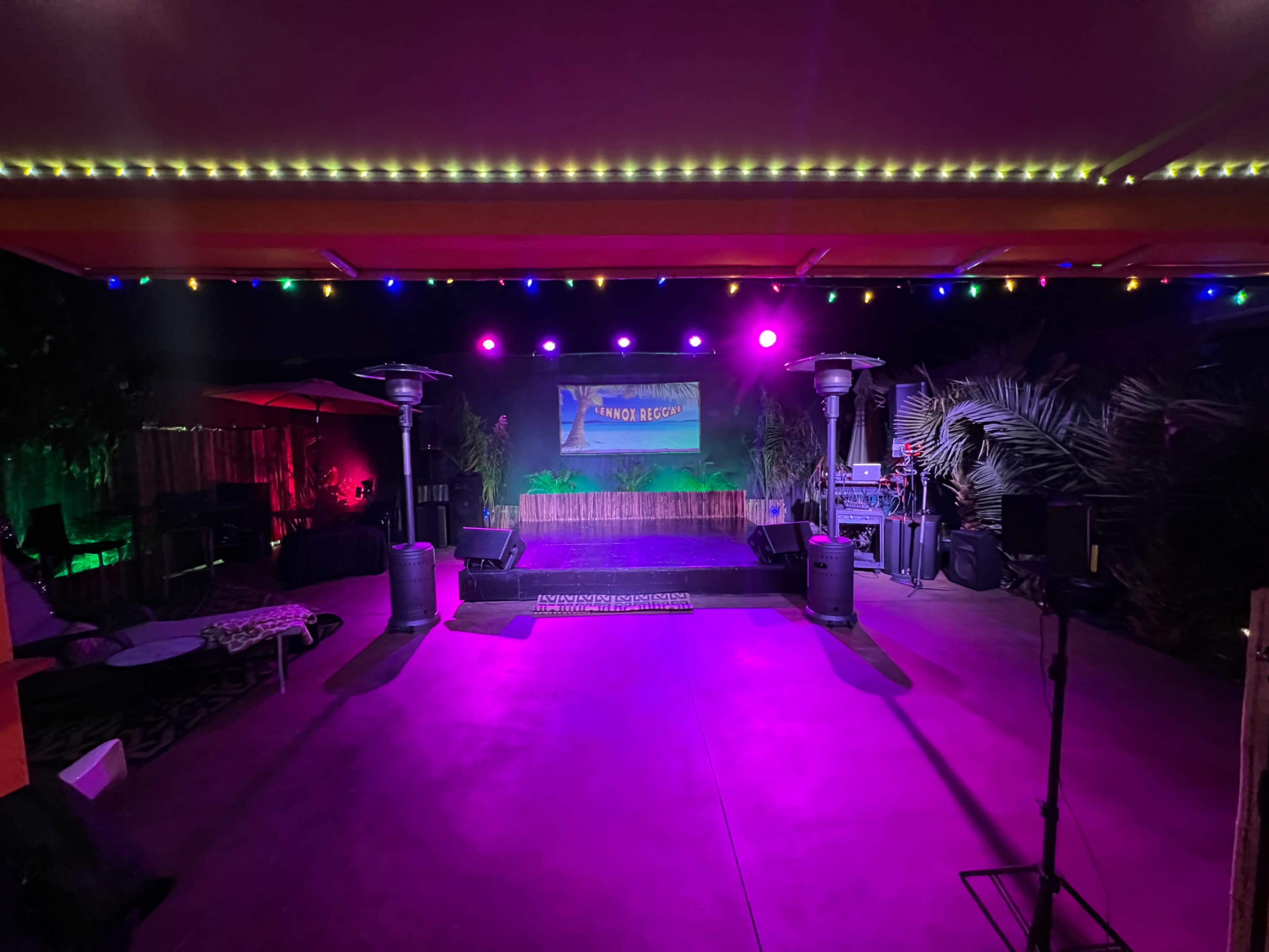 A stage set up outdoors with colorful lights, surrounded by decorative plants and equipment for a performance or event.