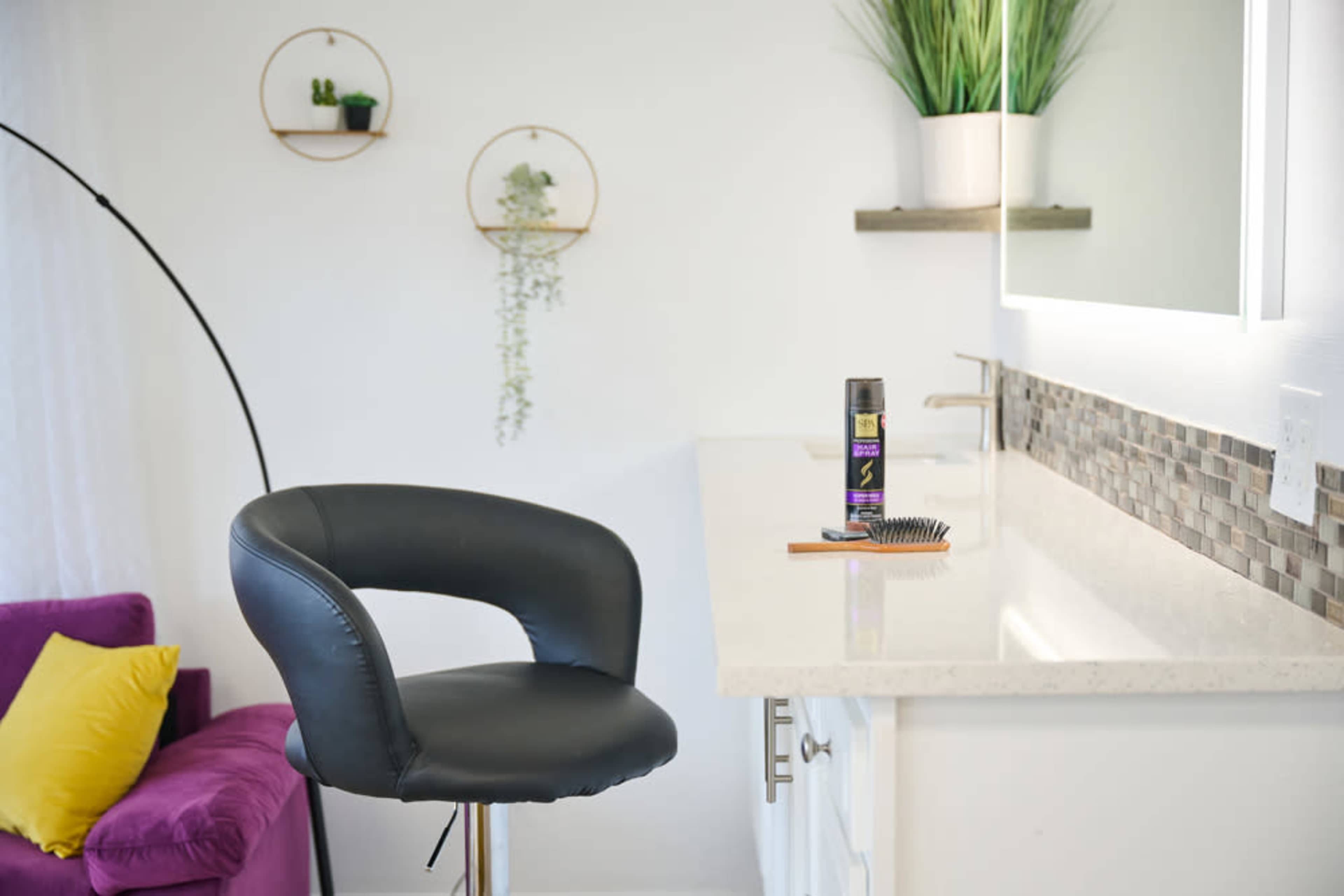 A modern vanity area features a black bar stool, a bottle of hair product, a comb, and decorative wall hangings with greenery.