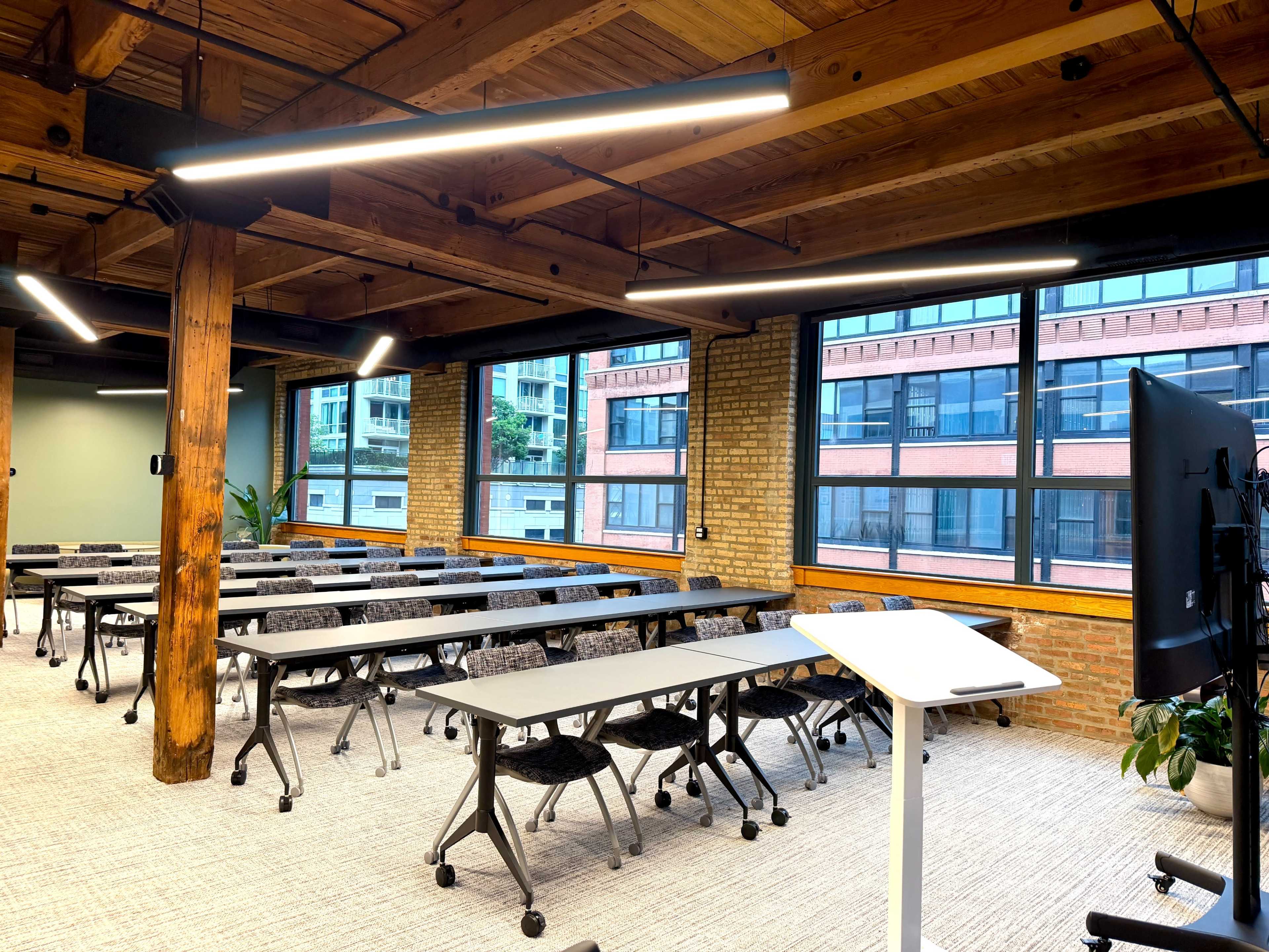 The image shows a modern conference room with rows of movable tables and chairs, large windows, and exposed wooden beams.