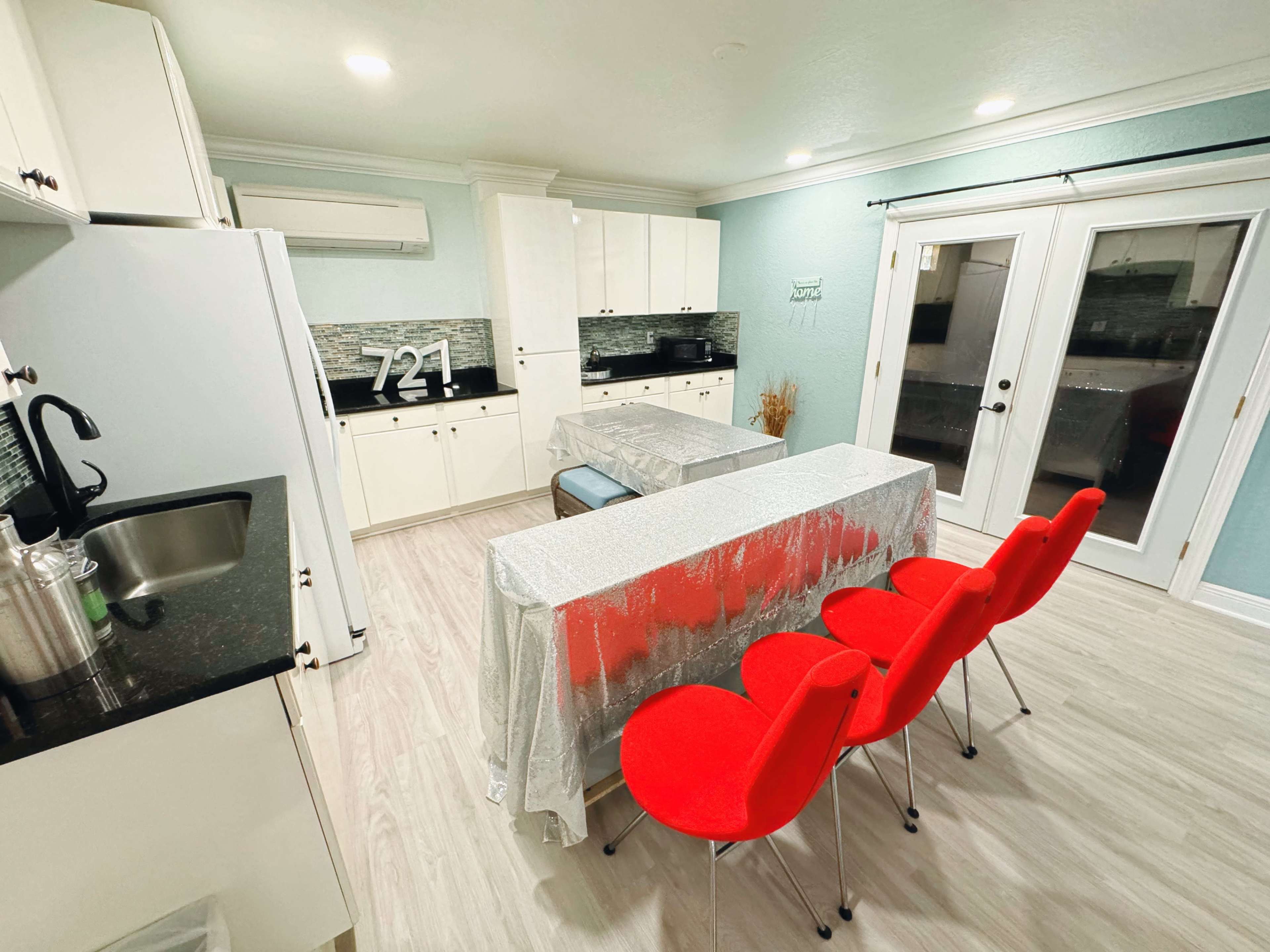 A modern kitchen features a black countertop, white cabinets, and a dining table covered with a silver tablecloth, accompanied by red chairs.