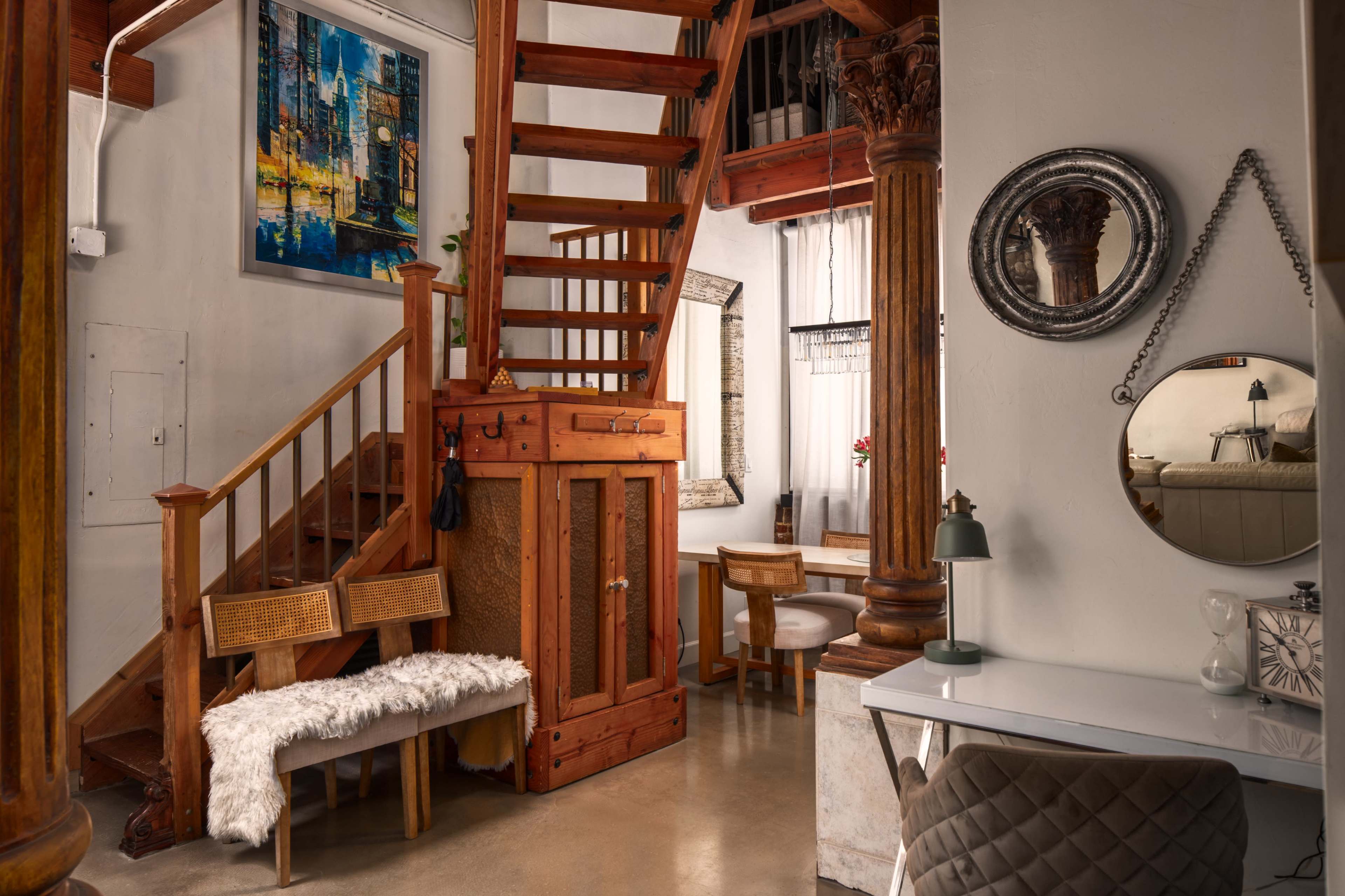 The image shows an interior space featuring a wooden staircase, a small seating area with a fur-like throw, an ornate mirror, and a desk near a window.
