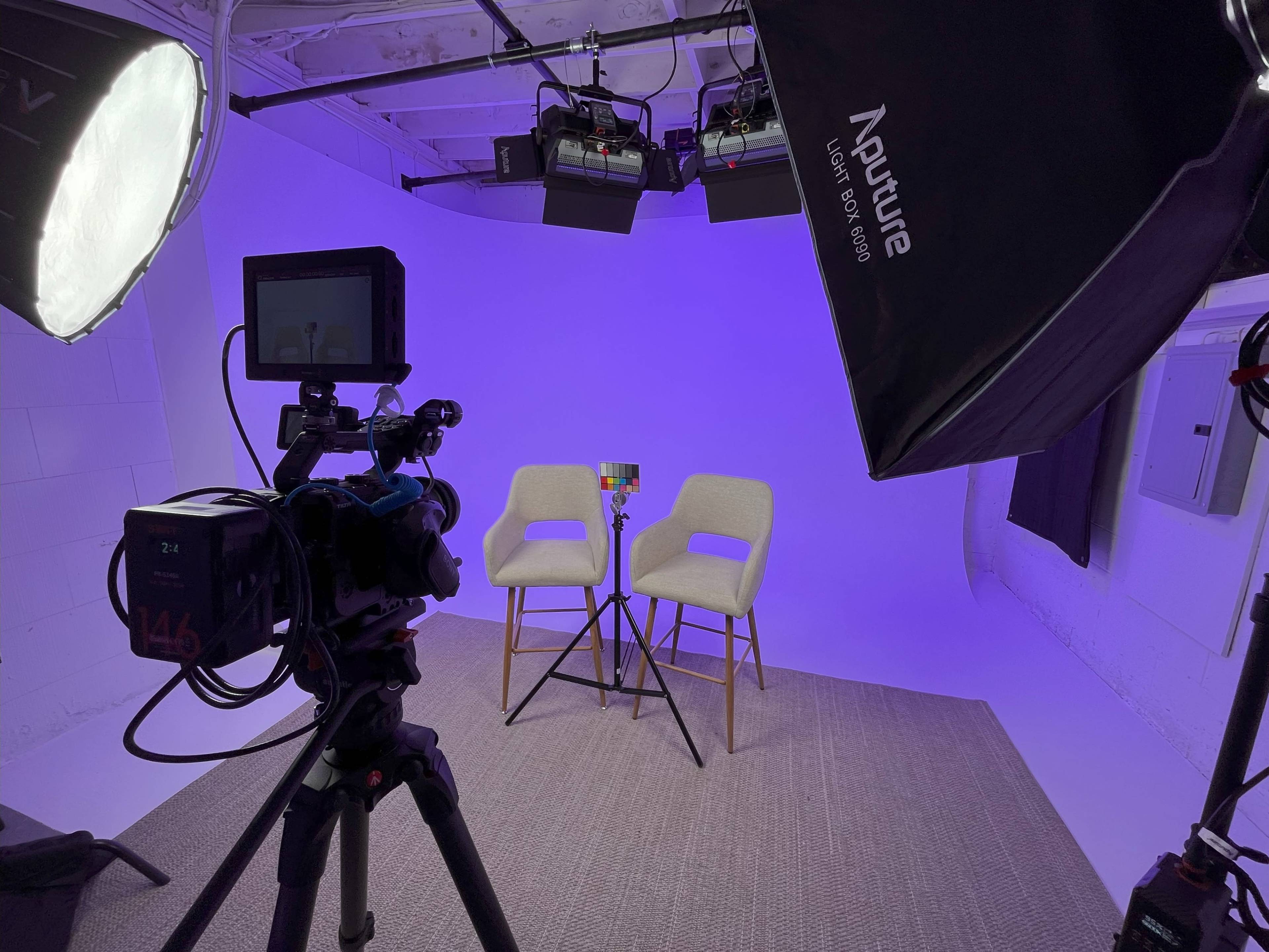 A recording studio setup with two chairs, a camera on a tripod, and studio lights against a purple backdrop.