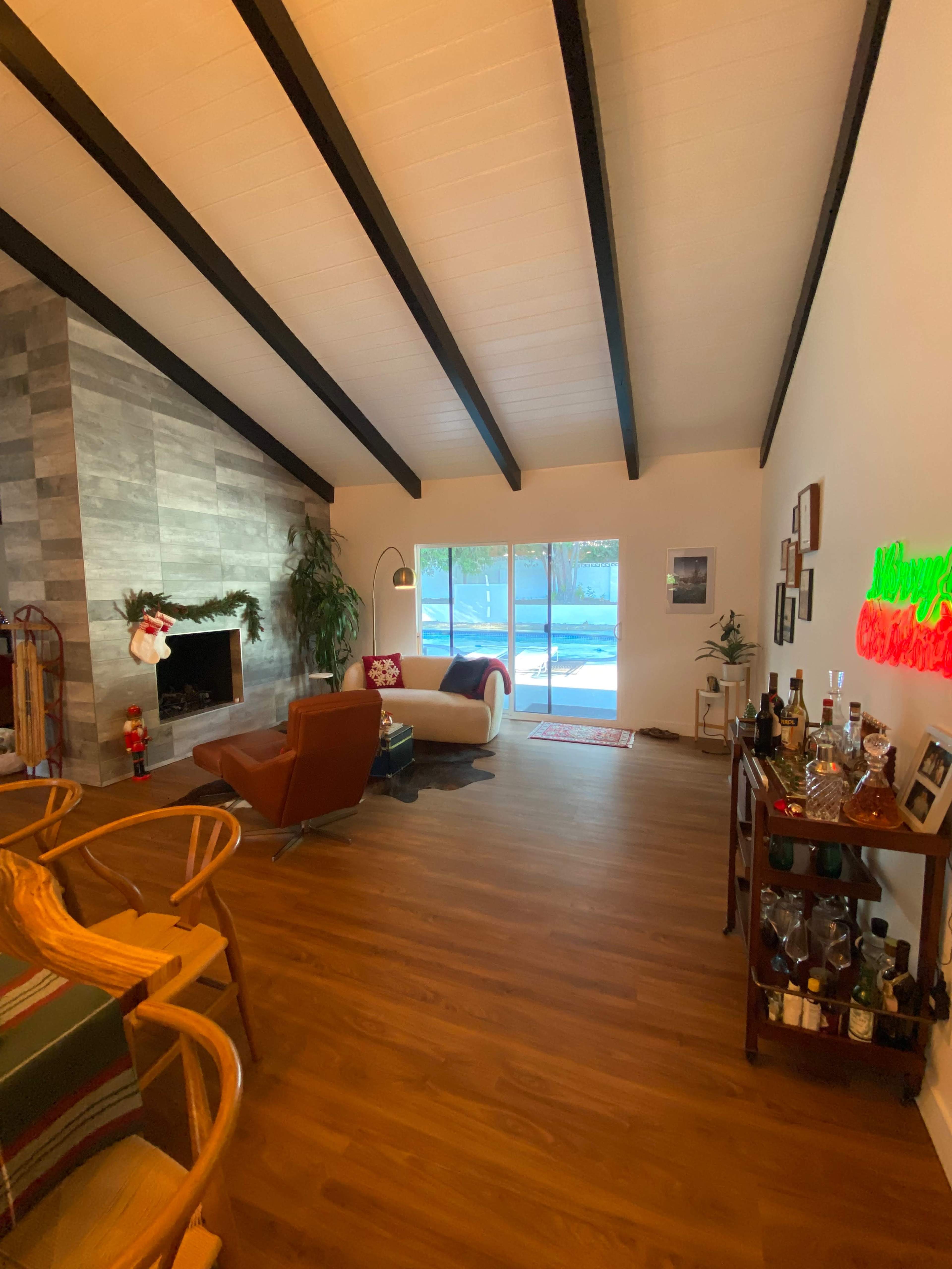 The image shows a spacious living room with a stone fireplace, wooden beams on the ceiling, and a view of a patio through large windows.