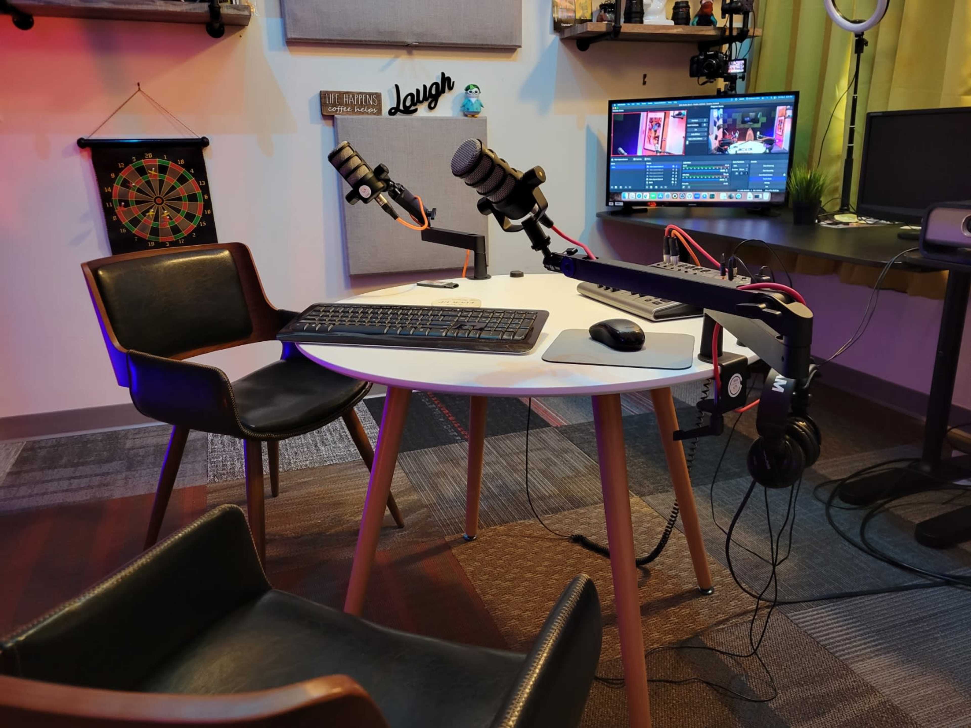 A desk setup featuring two microphones on adjustable arms, a keyboard, a headset, and a monitor displaying software, surrounded by two chairs.