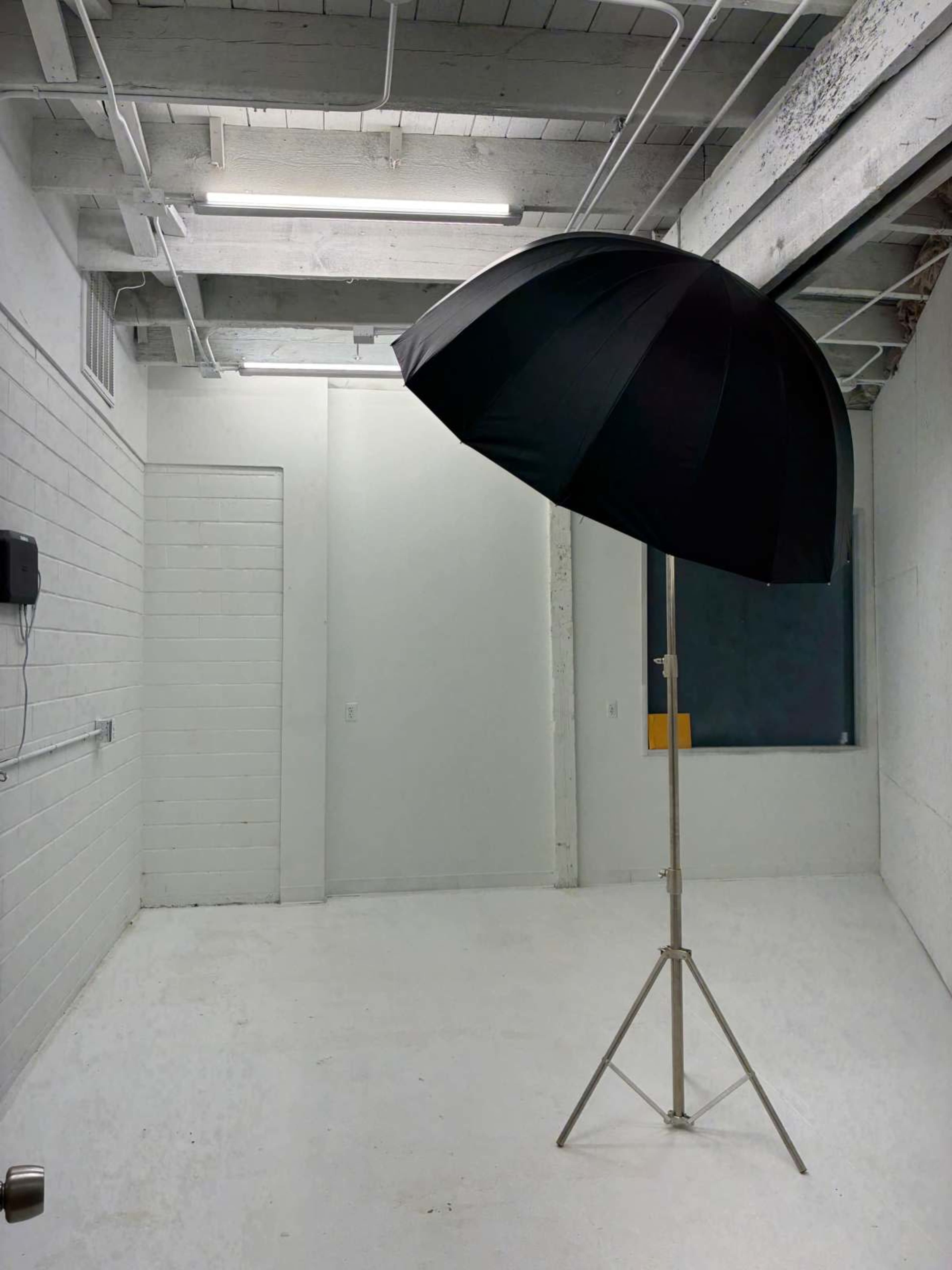 A large black umbrella is positioned on a stand in an empty, well-lit white room with concrete walls.