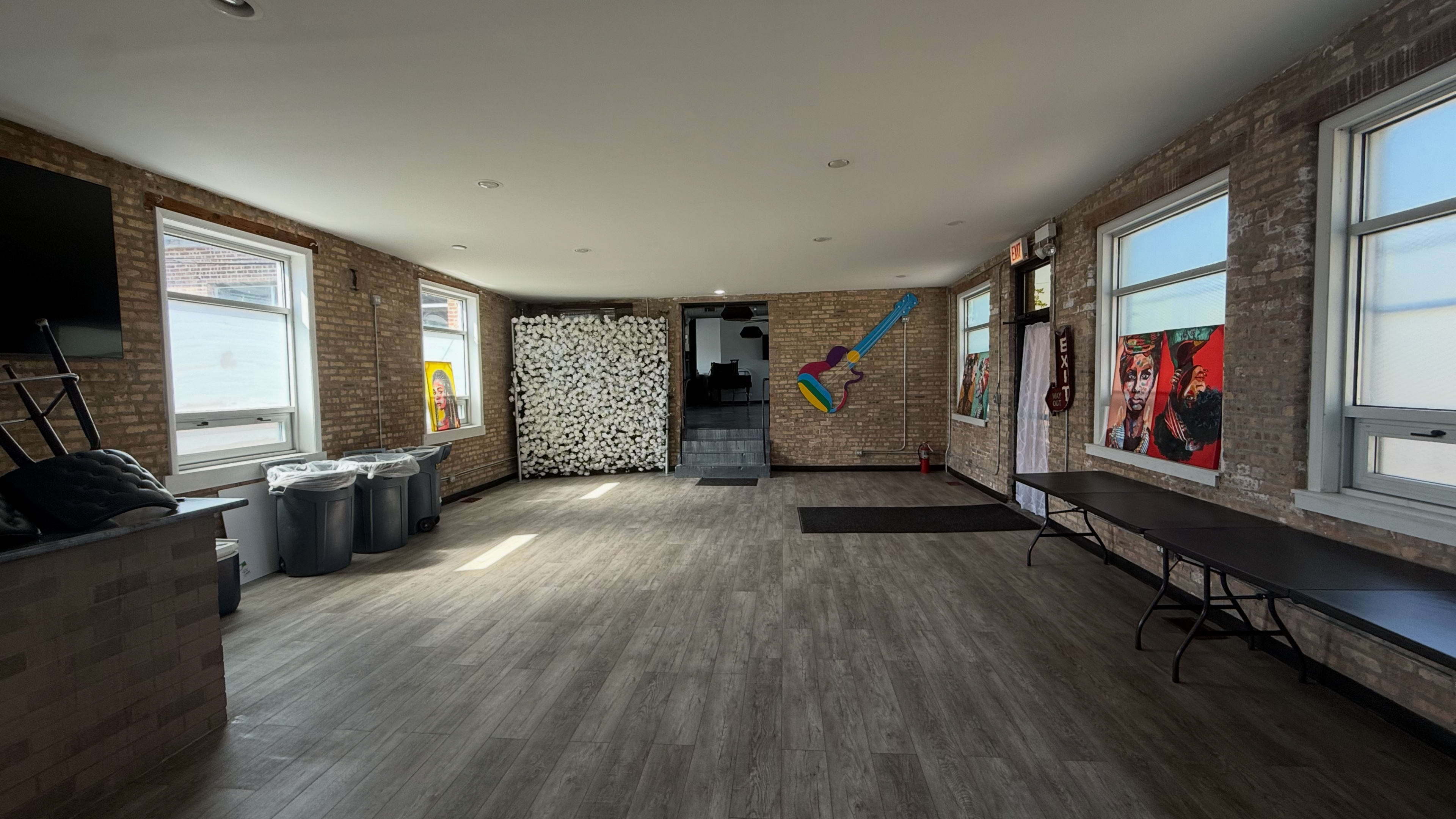 The image shows a spacious room with a wooden floor, featuring a wall decorated with artwork, a stack of chairs, and trash bins in the corner.