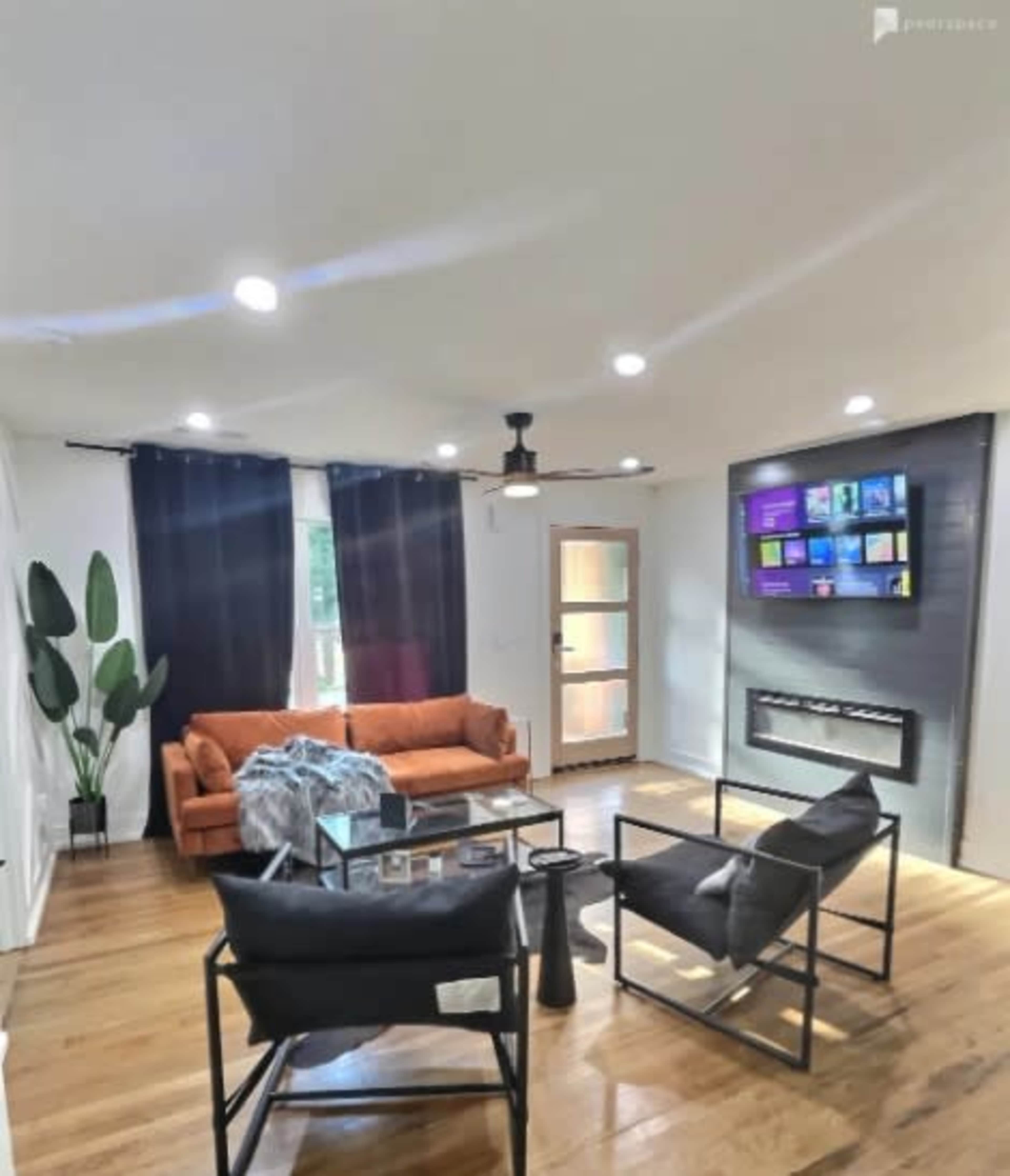 A modern living room features an orange sofa, black armchairs, a wall-mounted TV, and a fireplace, with large windows and hardwood floors.