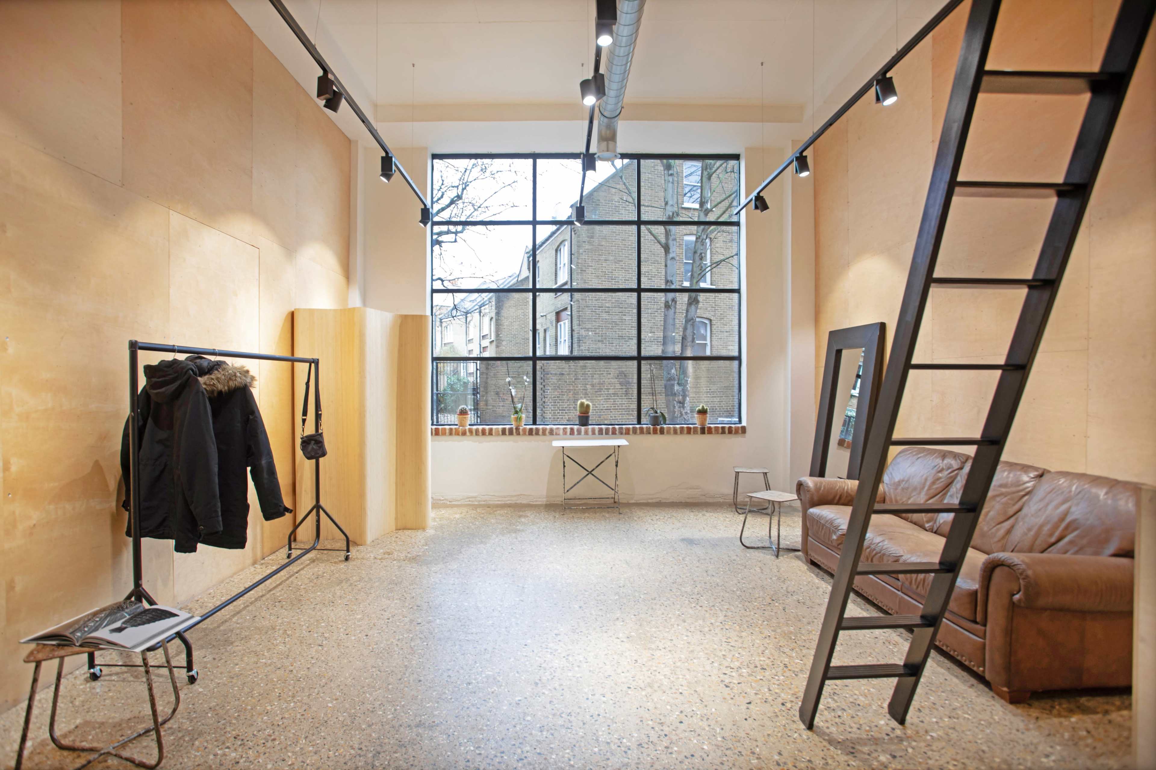 A spacious room with large windows, featuring a brown leather couch, a clothing rack with coats, a ladder, and a minimalist design.