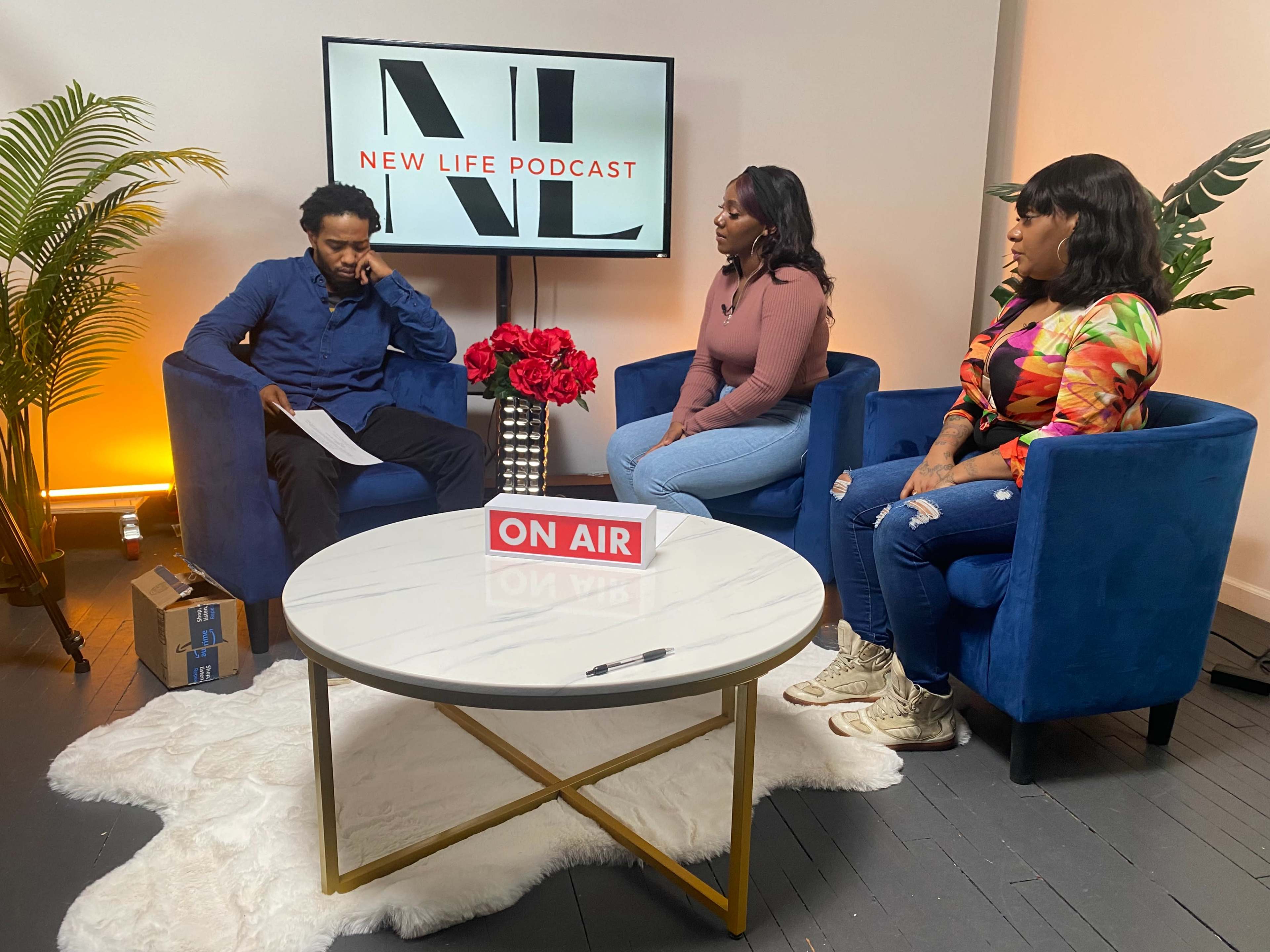 A podcast recording set features three participants seated in blue chairs around a white coffee table adorned with flowers and an "ON AIR" sign.