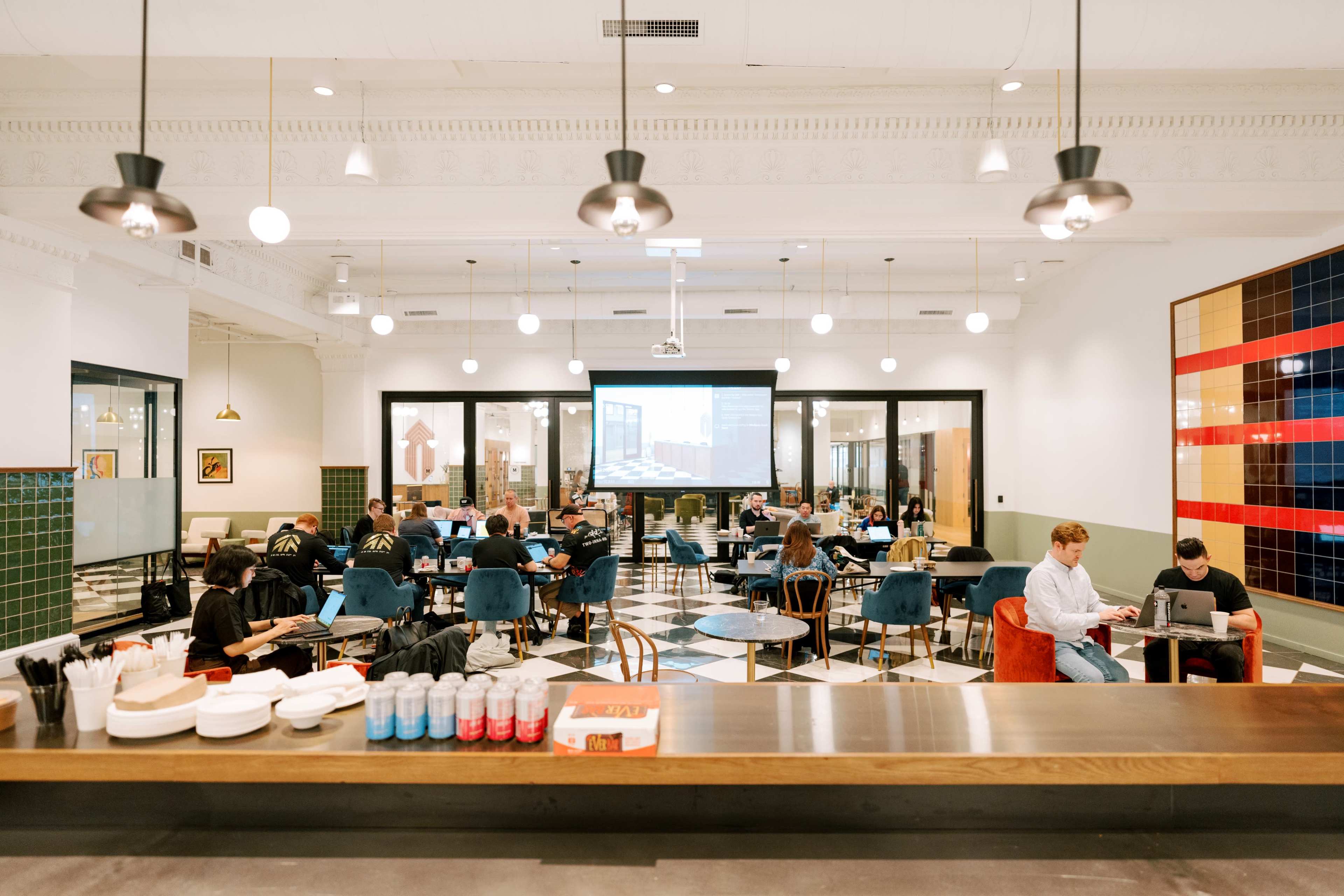 The image shows a spacious, modern café with various seating arrangements and people engaged in work and conversation, while a large screen displays content at the front of the room.