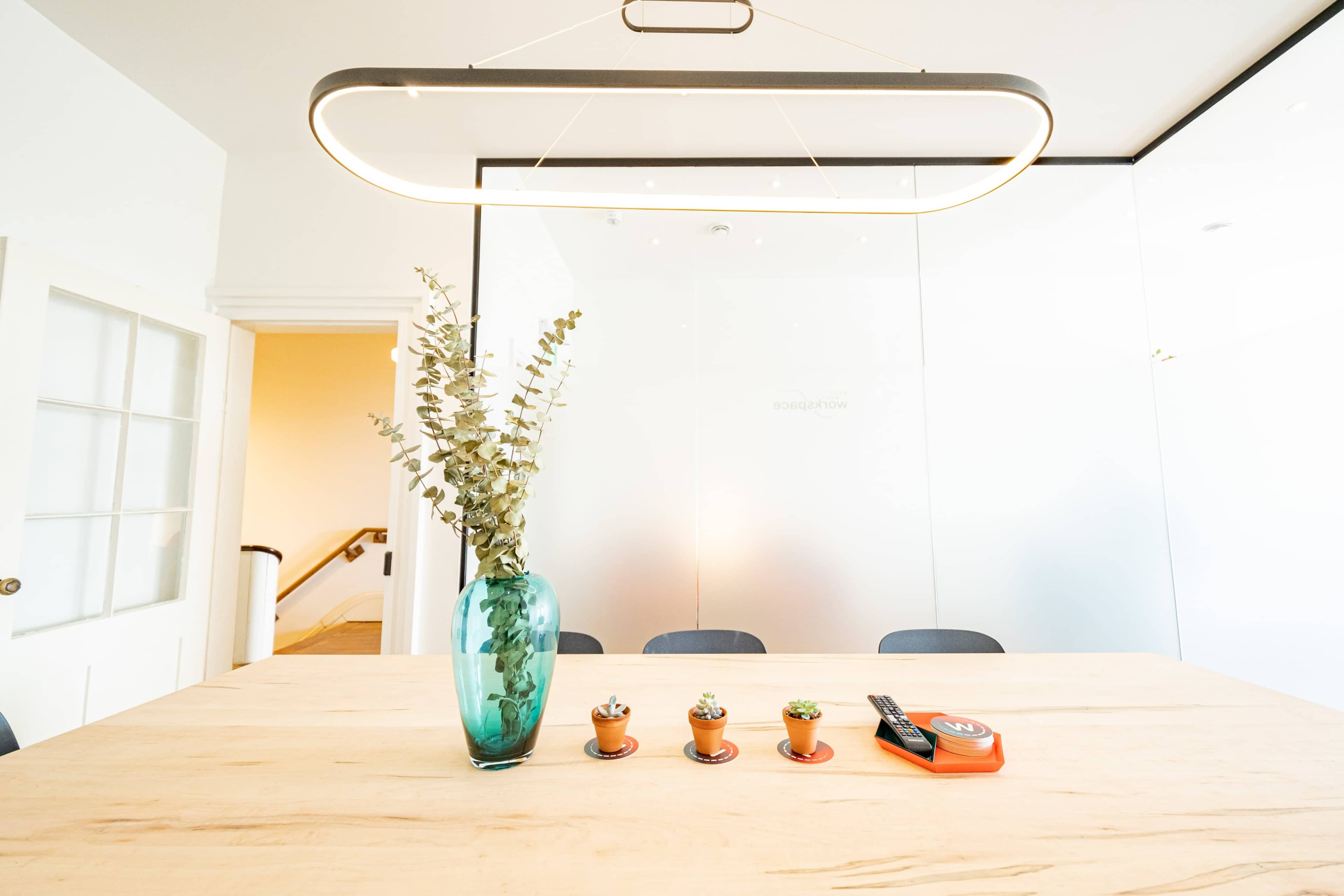 A modern conference room features a wooden table with a turquoise vase, small potted plants, and a measuring tape under a hanging light fixture.