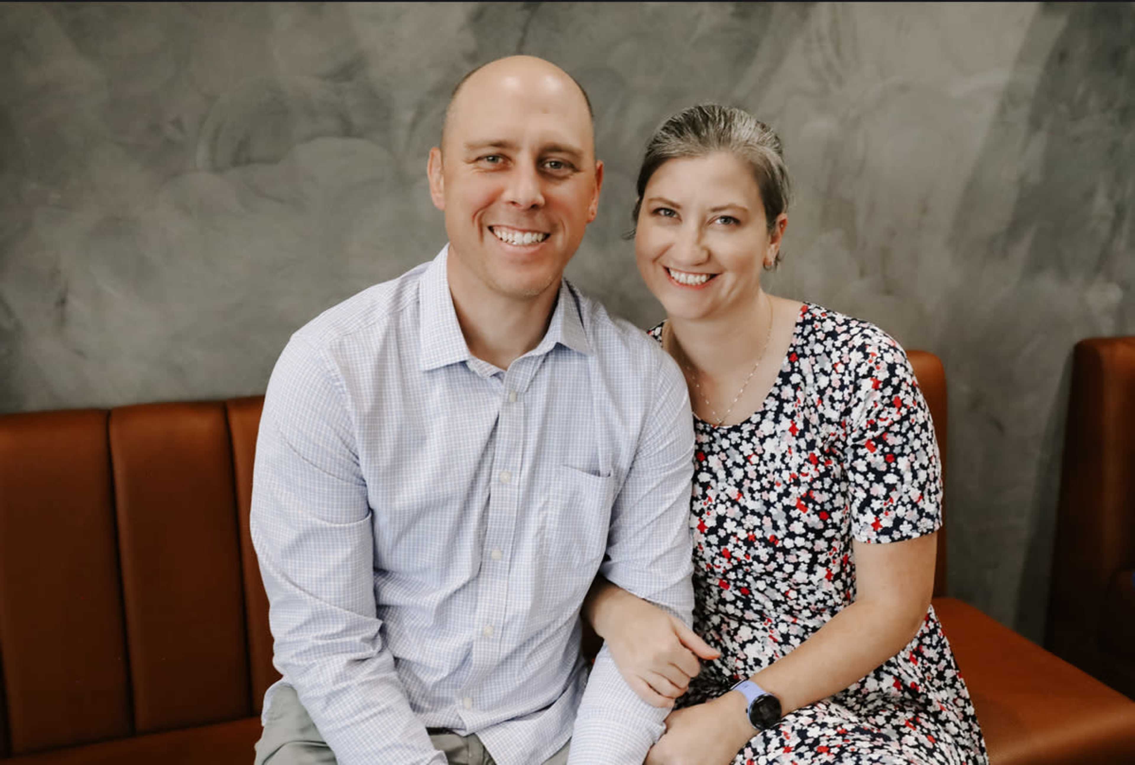 A man and a woman sit close together on a brown couch, smiling at the camera.
