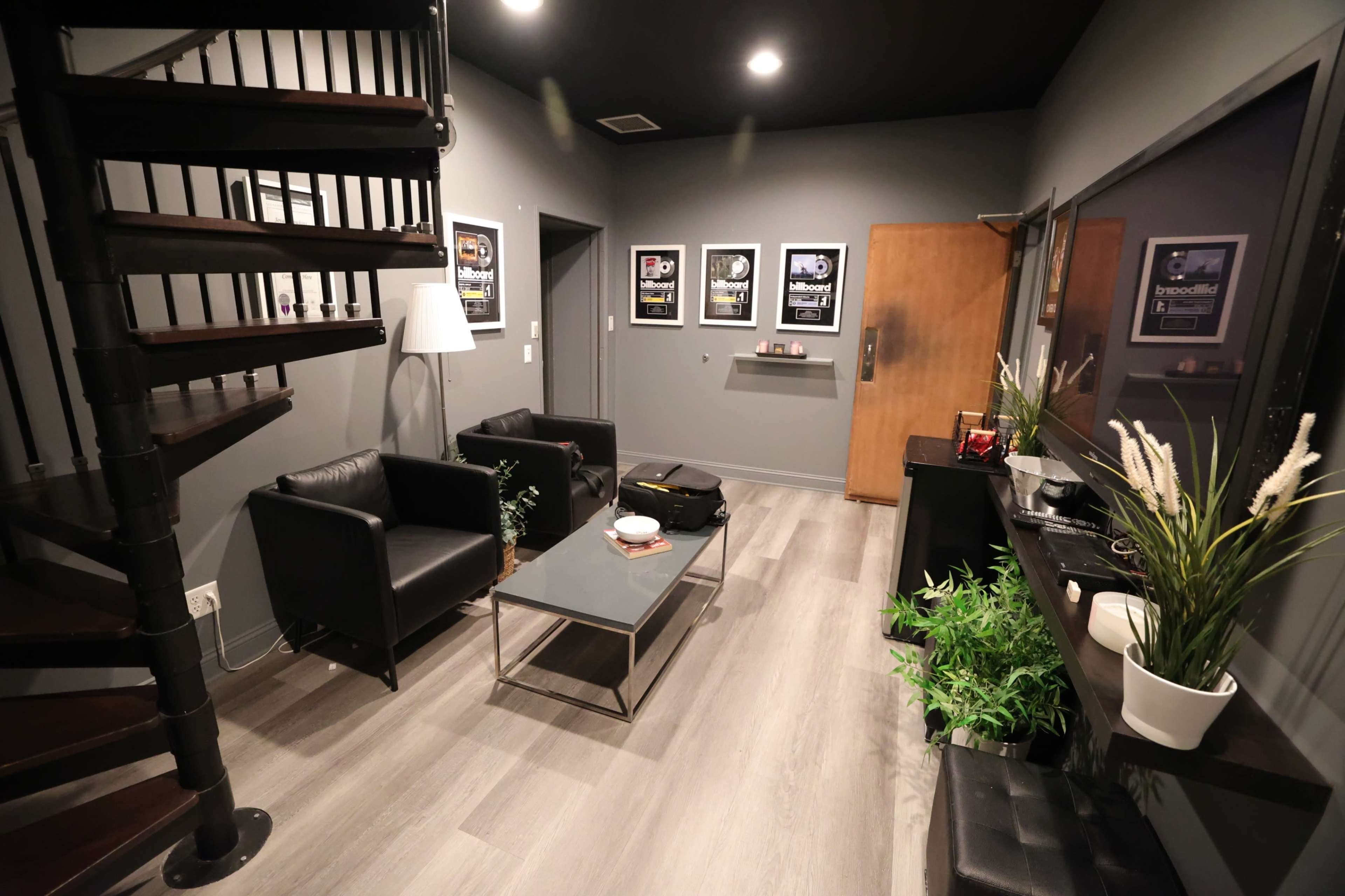 The image shows a modern, minimalistic room with a spiral staircase, two black armchairs, a glass coffee table, and several framed posters on the gray walls.