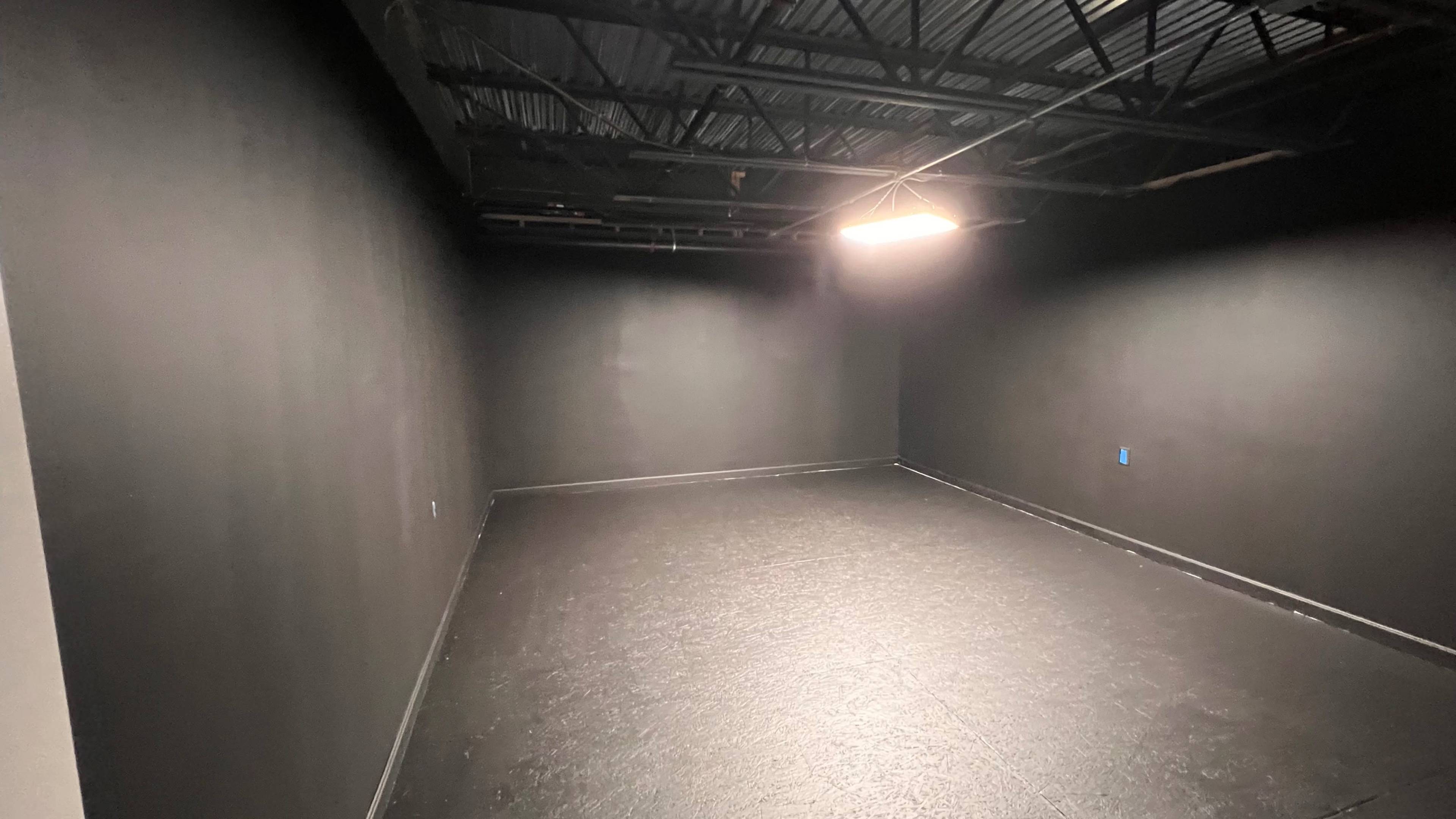 A sparsely furnished black-walled room with a black floor and a single overhead light fixture.