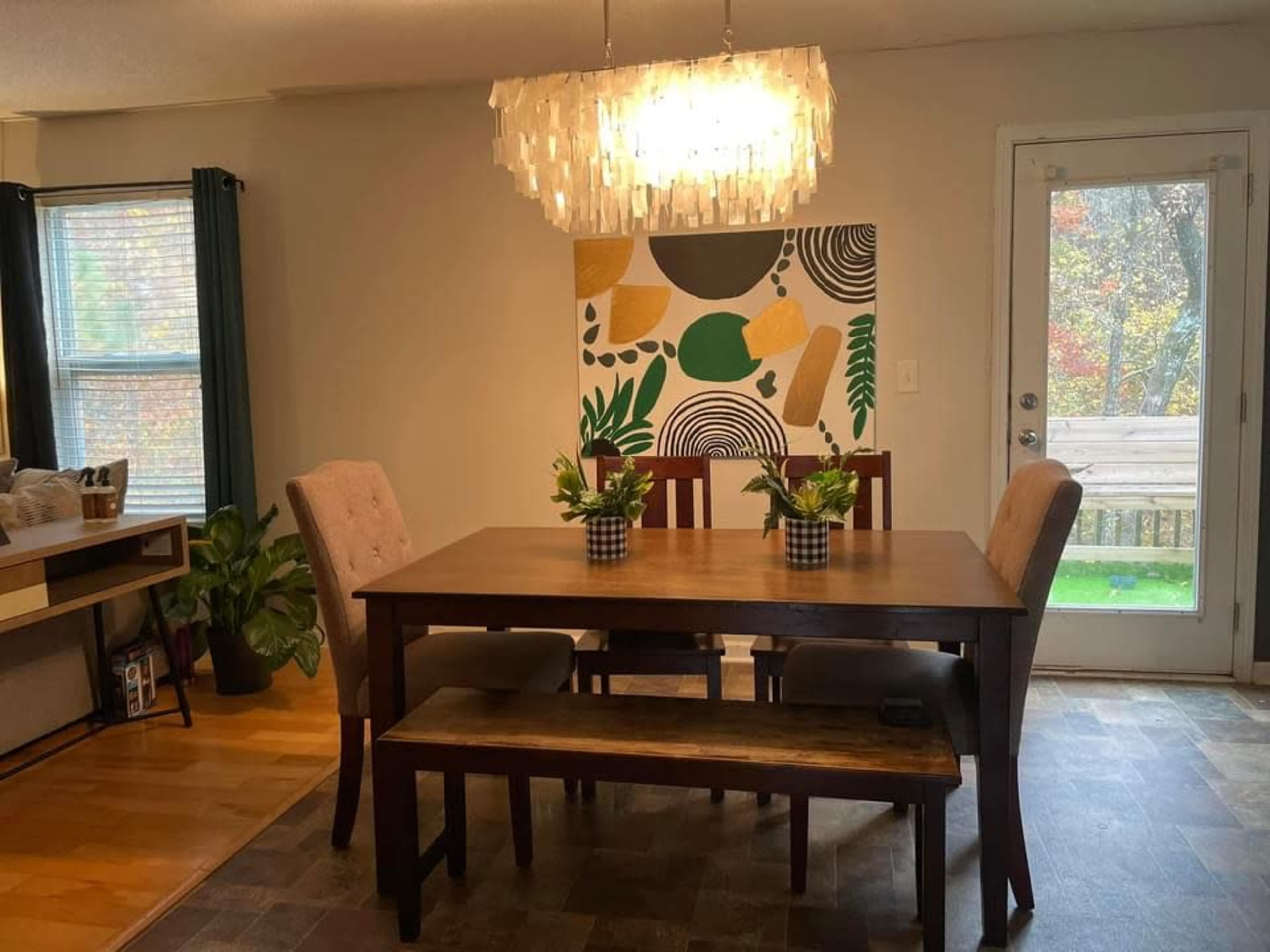 A dining area features a wooden table surrounded by four chairs and a bench, with a colorful wall art piece and a chandelier overhead.