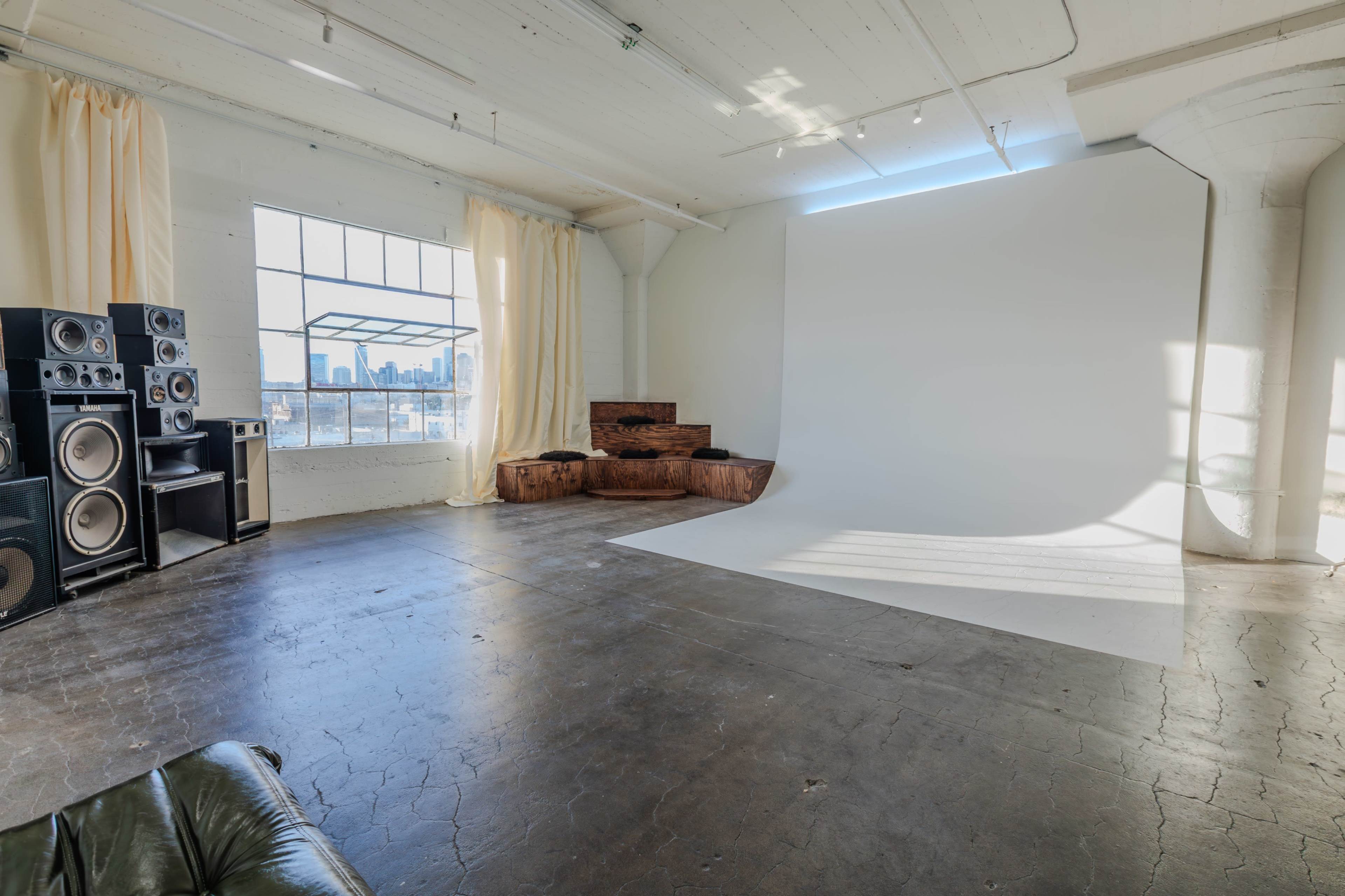 The image shows a spacious room with a blank white backdrop, a black couch, large speakers, and wooden blocks arranged as seating, illuminated by natural light from a nearby window.