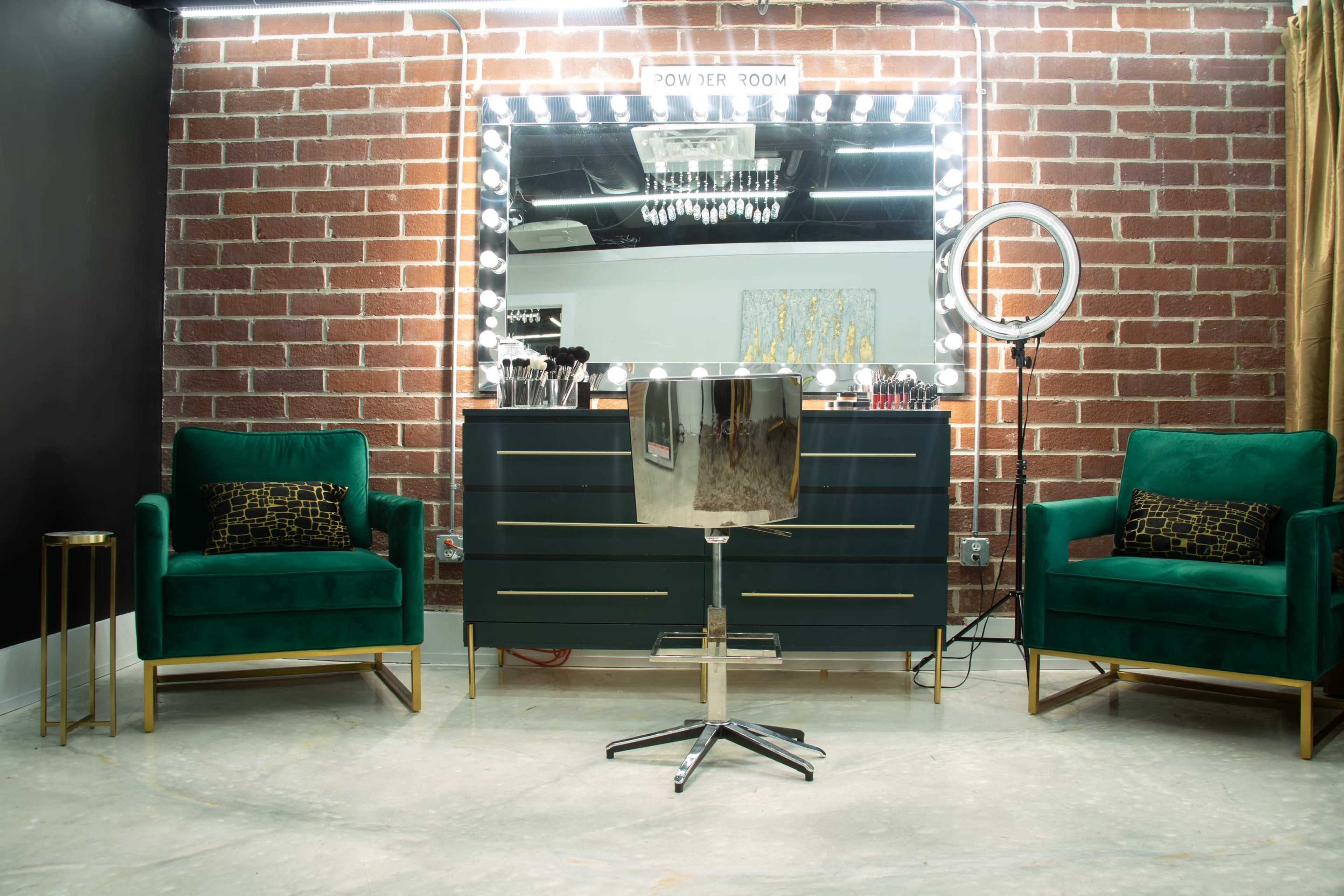 A stylish beauty setup featuring a large mirror with lights above it, a metallic salon chair in front, and two green velvet armchairs on either side, all set against a brick wall.