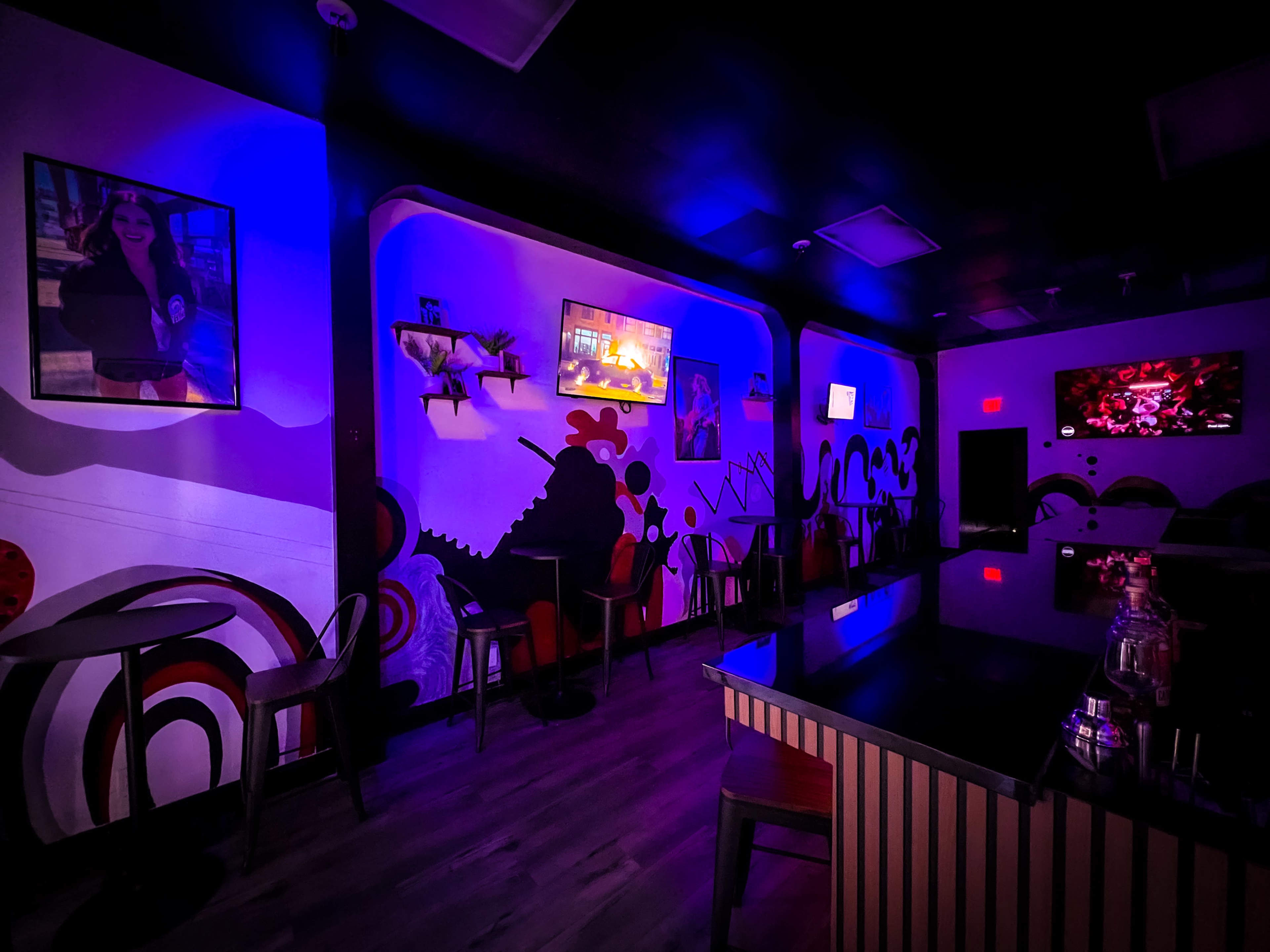 A dimly lit room with vibrant murals on the walls and a bar counter, featuring several screens displaying images.