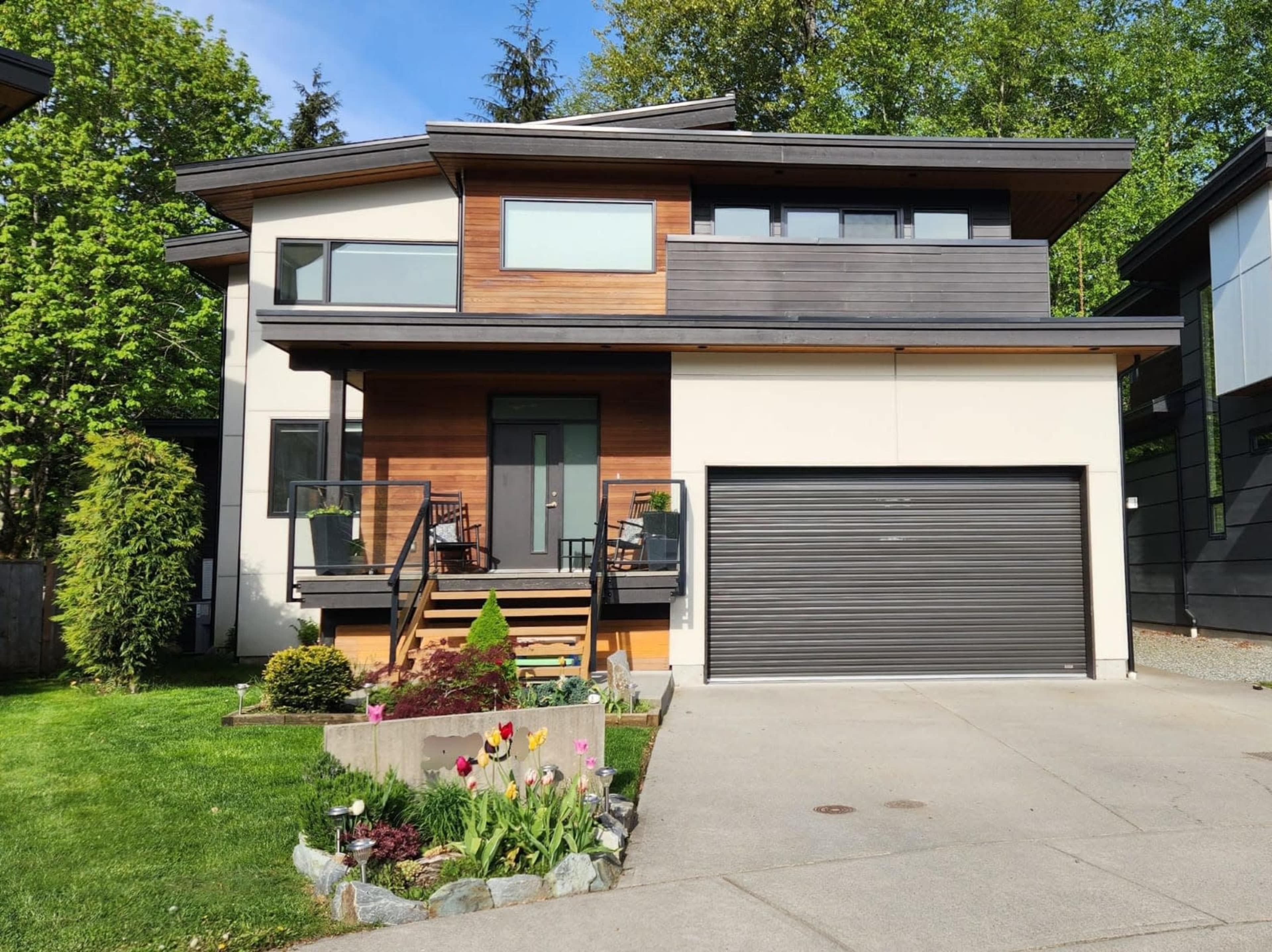 A modern two-story house with a wooden facade and a landscaped front yard featuring flowers and a paved driveway.