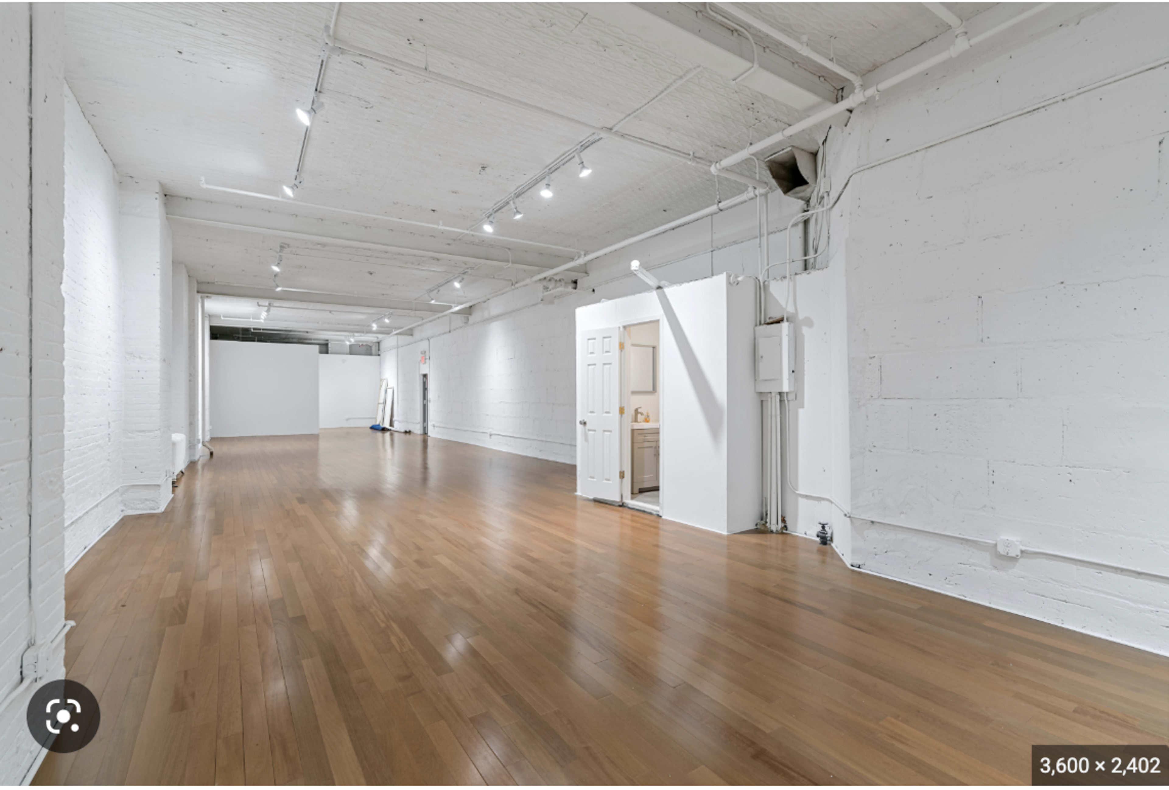 A spacious, empty interior room with wooden floors, white walls, and several overhead lights.