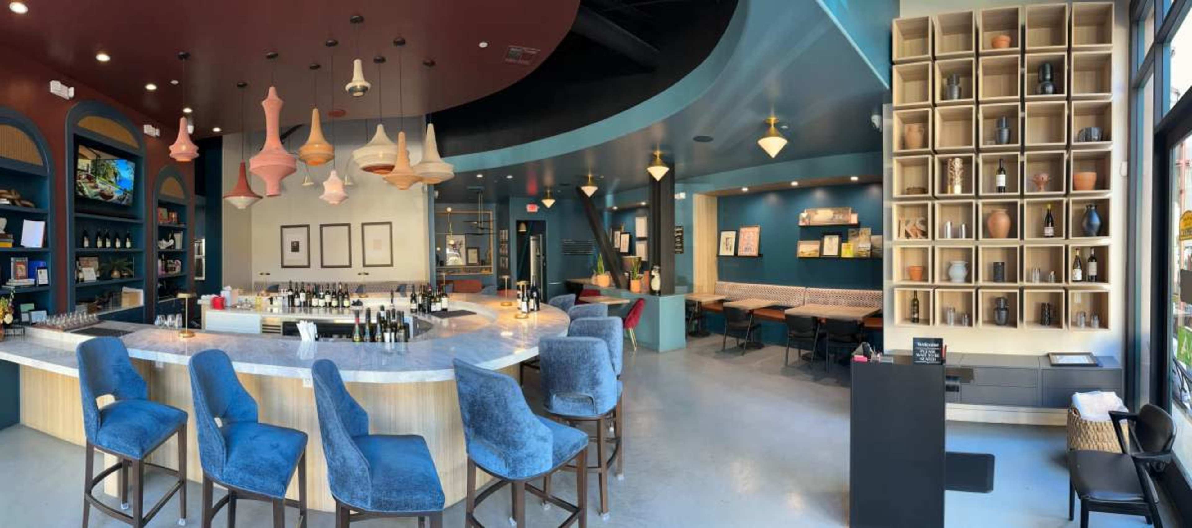 The image shows a modern wine bar featuring a curved counter with blue upholstered stools, a seating area with beige benches, and a wall displaying shelves of bottles and decorative items.