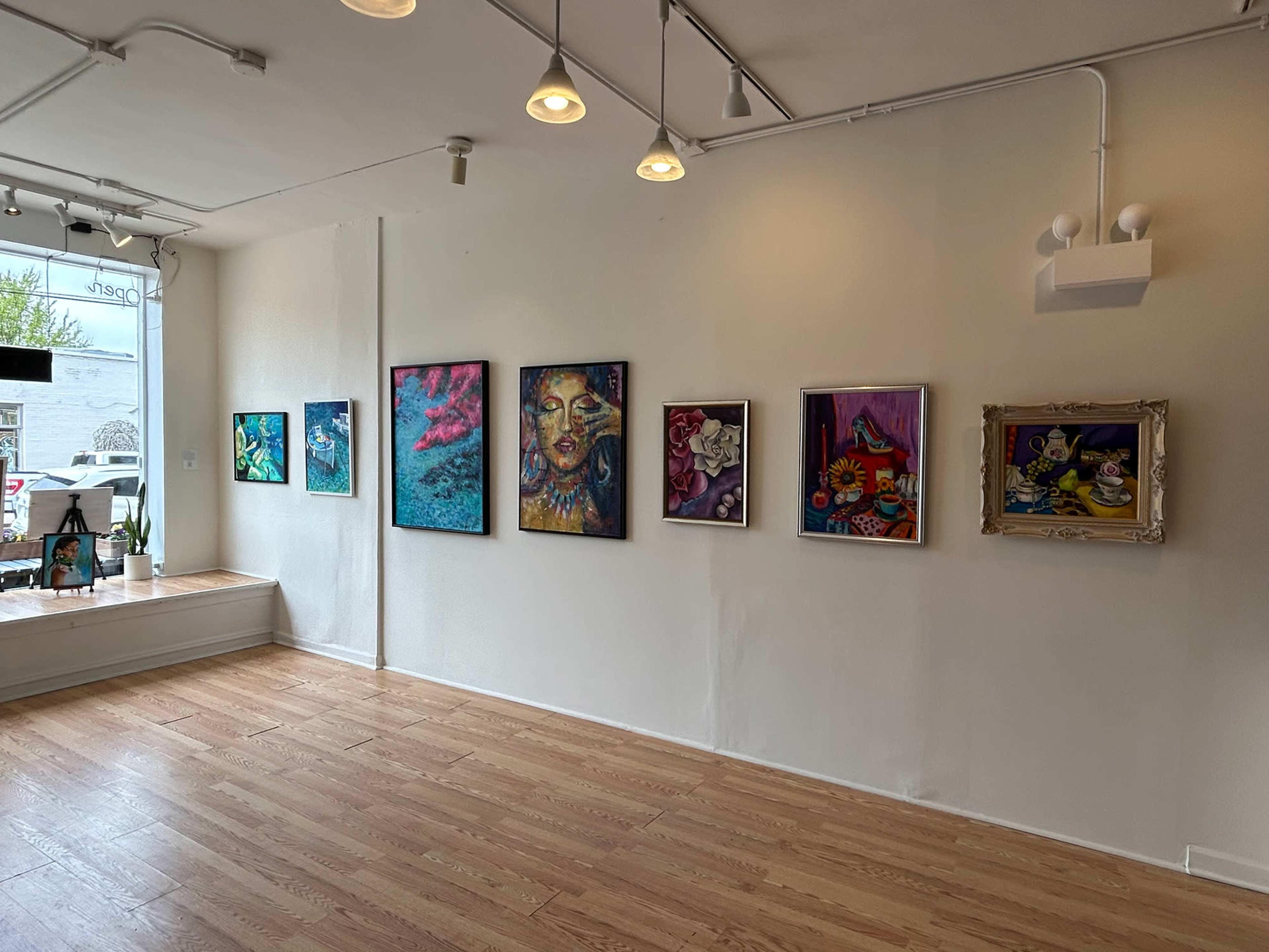 The image shows an art gallery interior featuring a series of framed paintings displayed on a white wall, with wooden flooring and natural light coming in from windows.