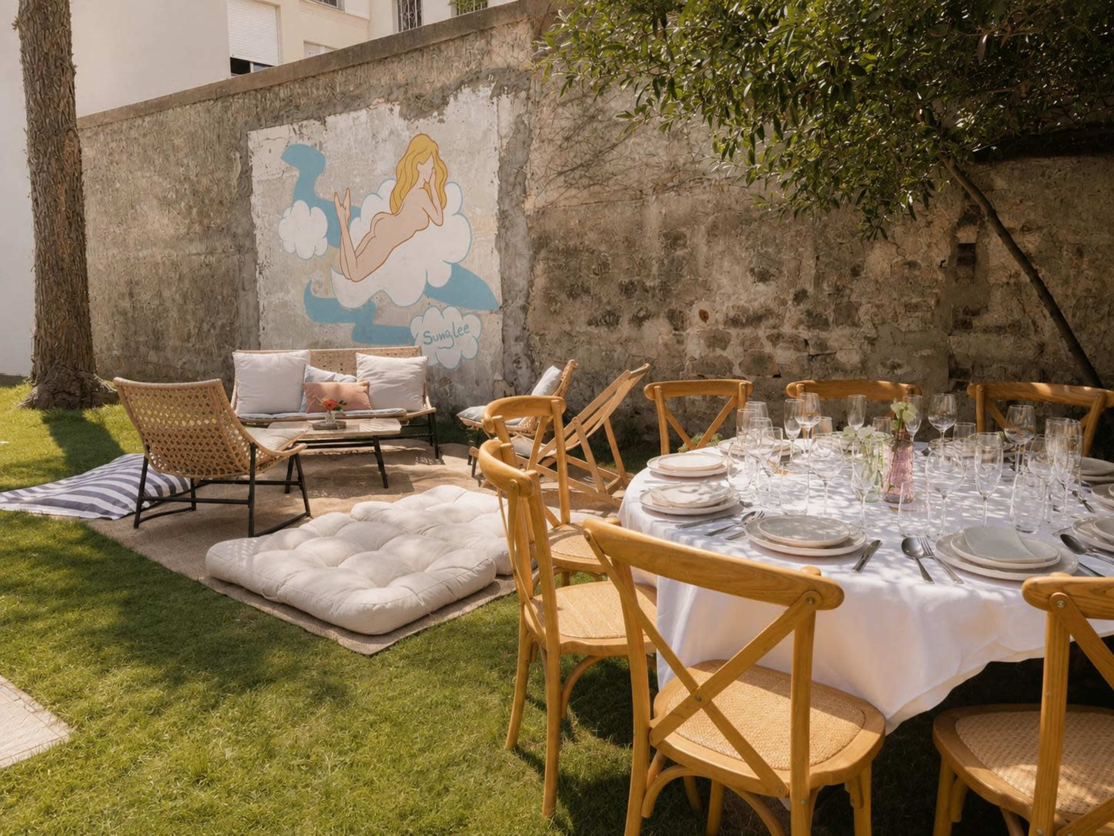A dining setup with a circular table surrounded by wooden chairs is located on a grass lawn, next to a seating area featuring a sofa and a mural on the wall.