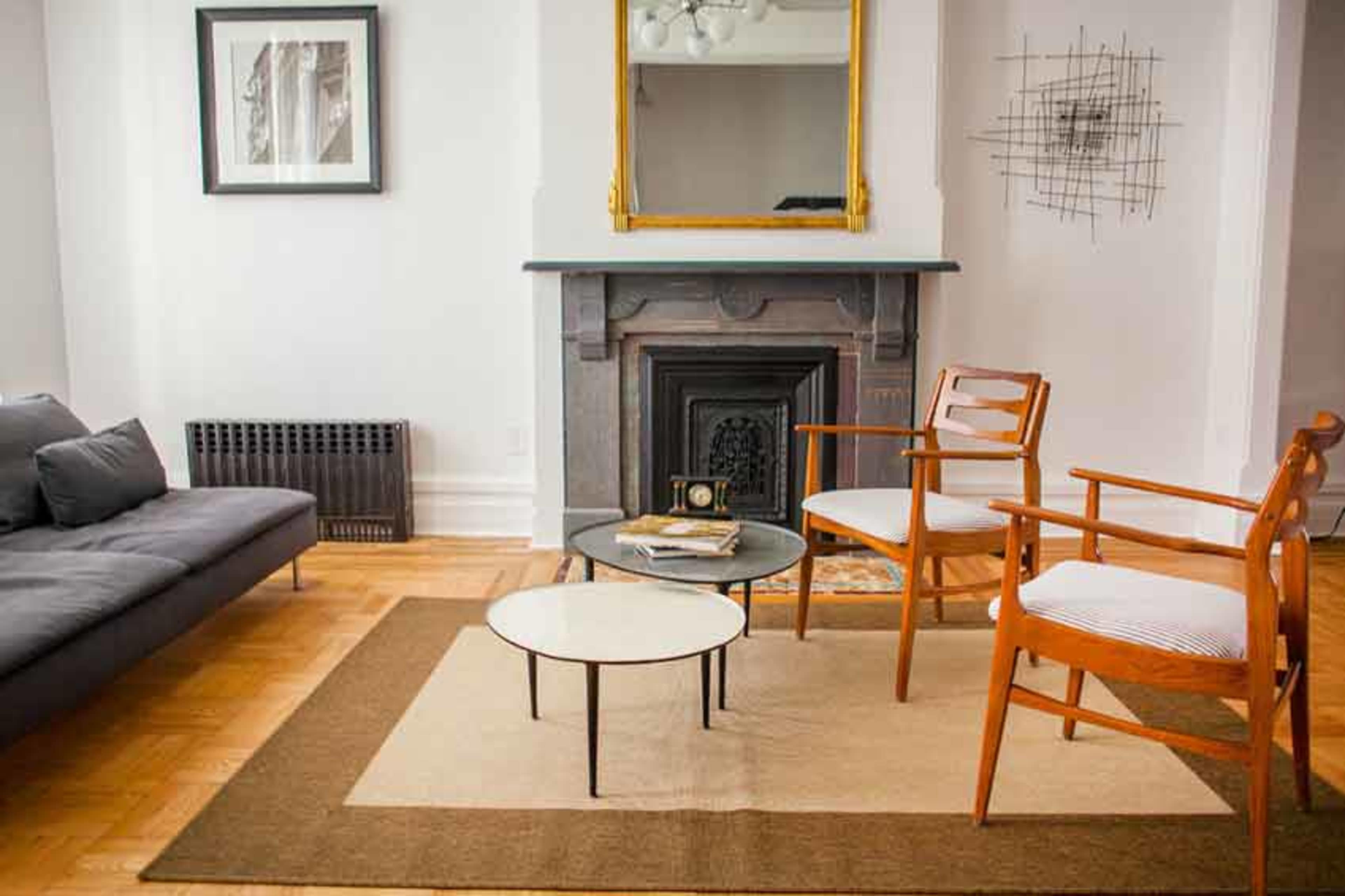 A living room features a gray sofa, two wooden chairs, a circular coffee table, and a fireplace with a mirror above it.