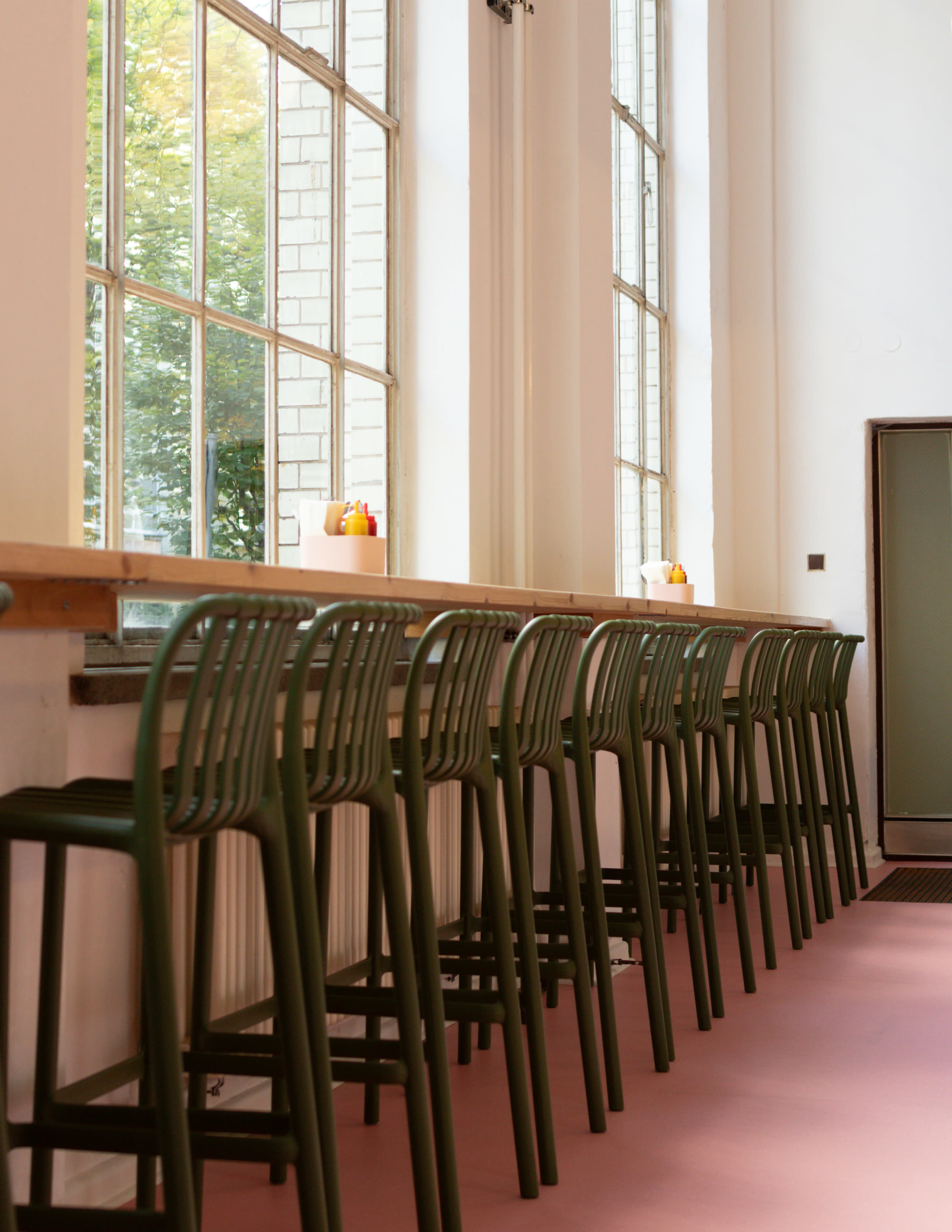 The image shows a row of green barstools lined up at a long wooden counter beside large windows.
