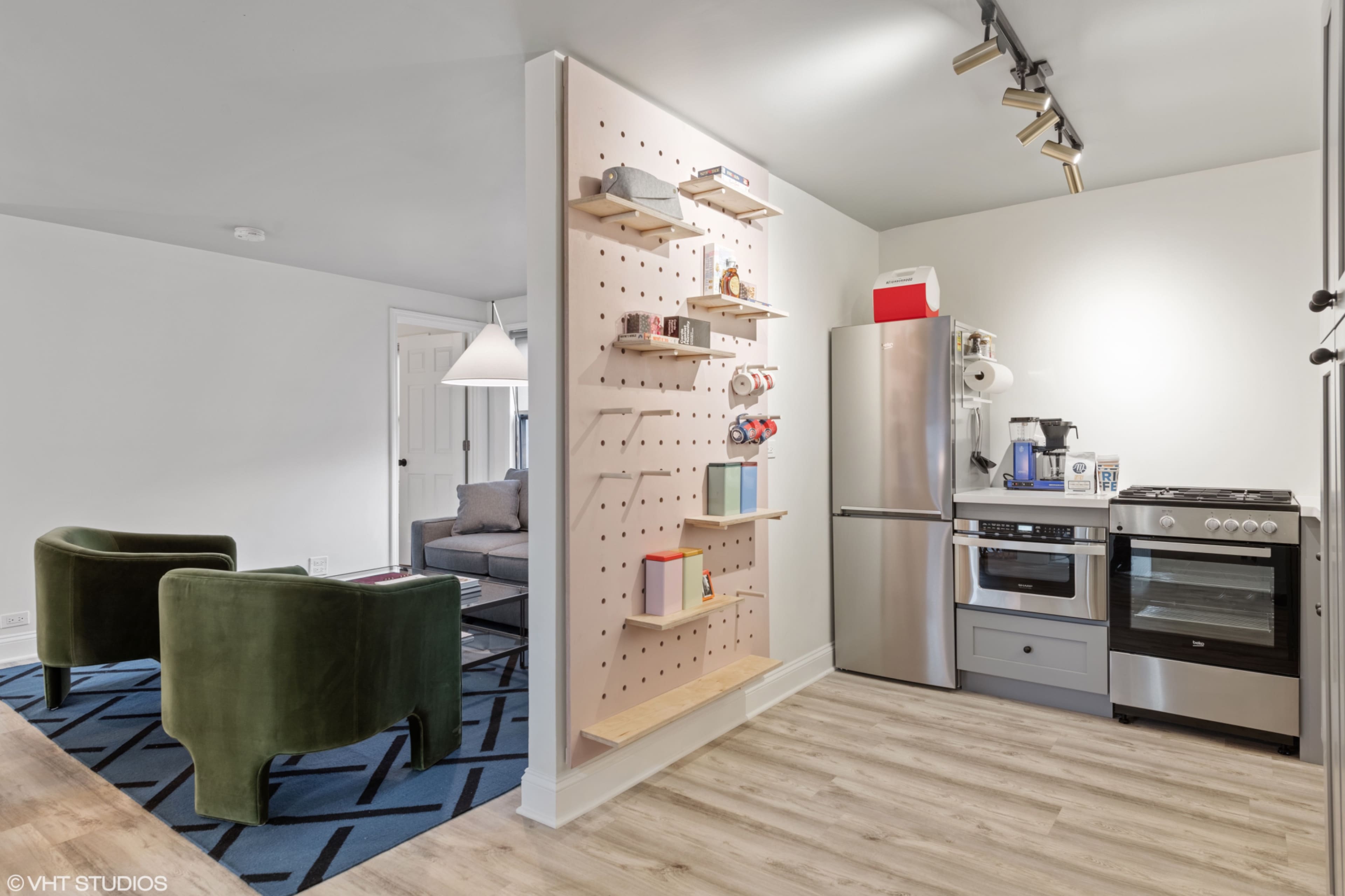 The image shows a modern kitchen area with stainless steel appliances and a wall featuring pegboard shelves for organizing items, alongside a cozy seating space with green chairs.