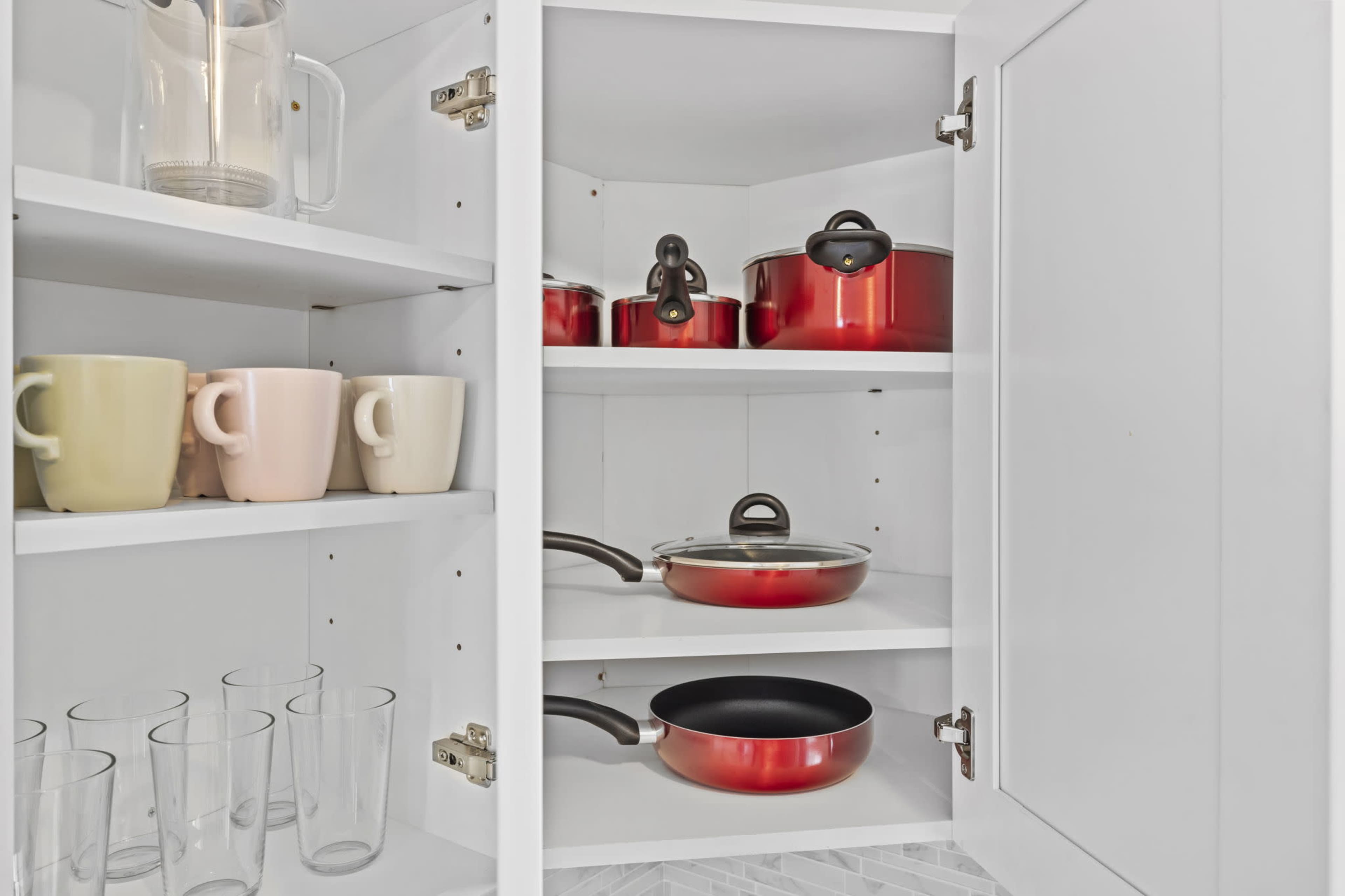 A white kitchen cabinet is open to reveal shelves containing red cooking pots, a frying pan, glasses, and mugs.