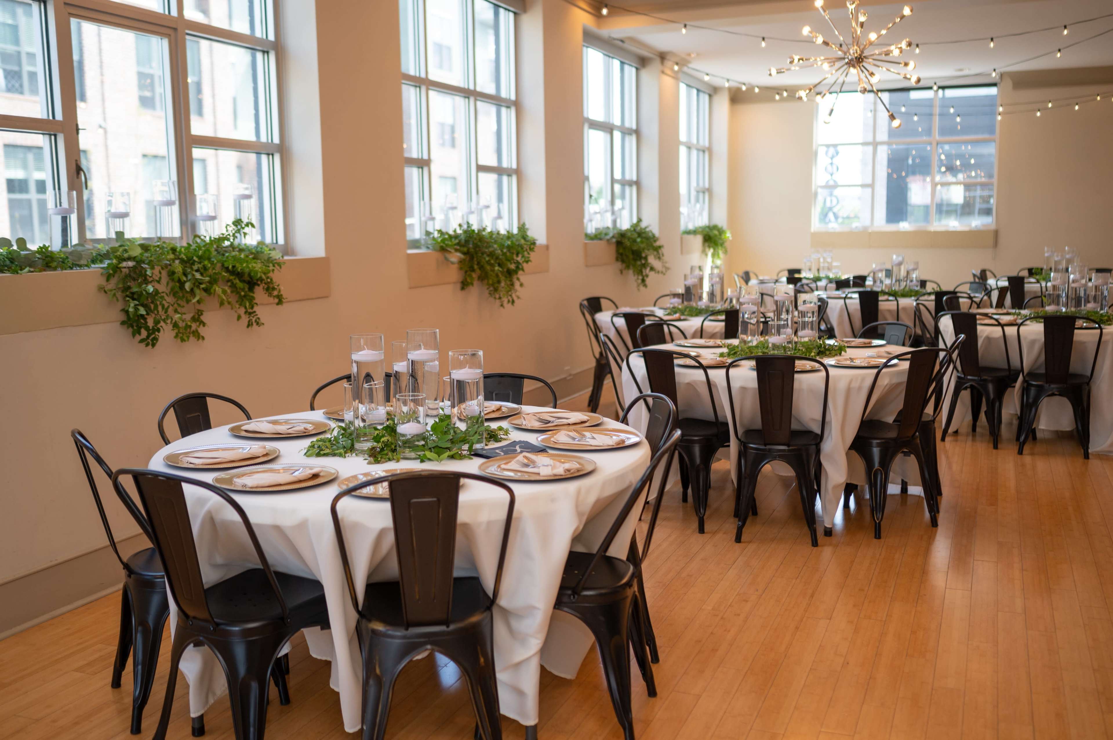 A banquet hall features round tables set with white tablecloths and black chairs, decorated with green foliage and glass centerpieces, illuminated by natural light through large windows.