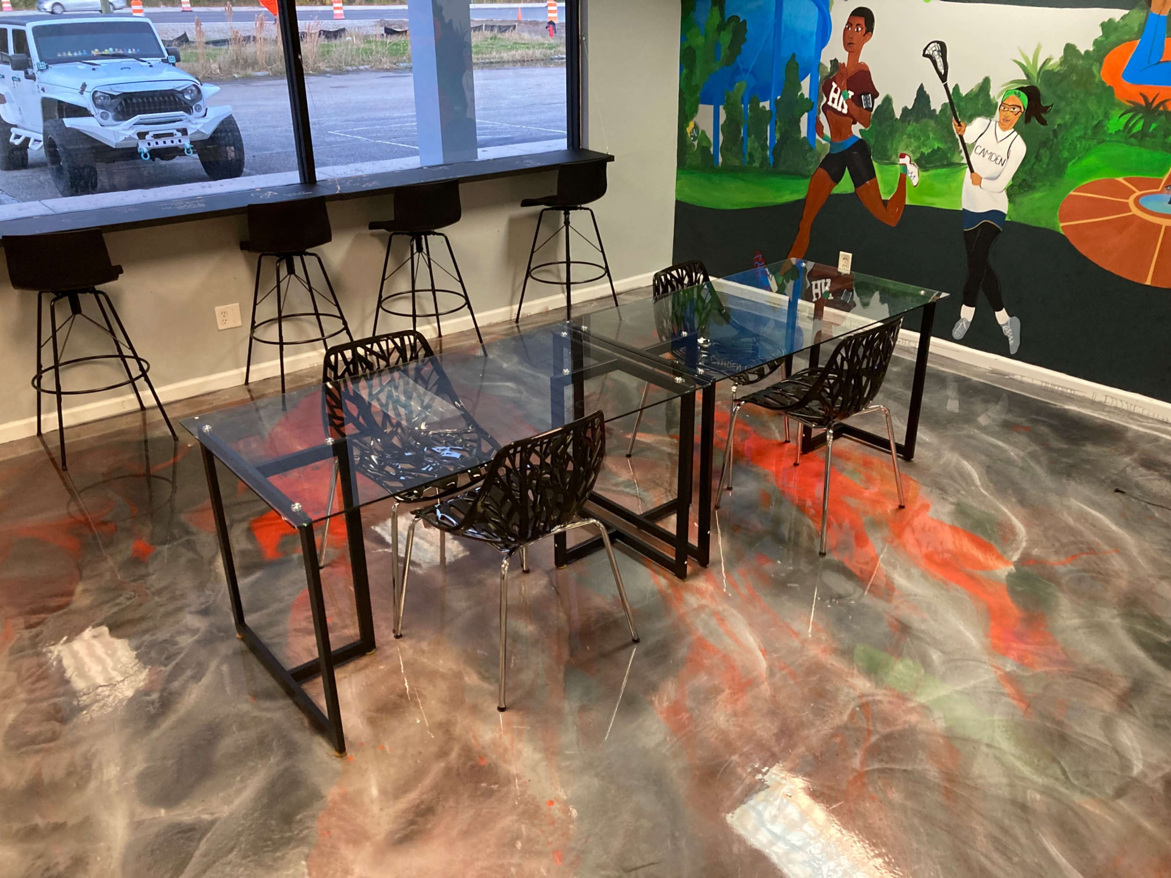 A modern office space features a glass-top dining table surrounded by black chairs, with high stools against the wall and a colorful mural in the background.