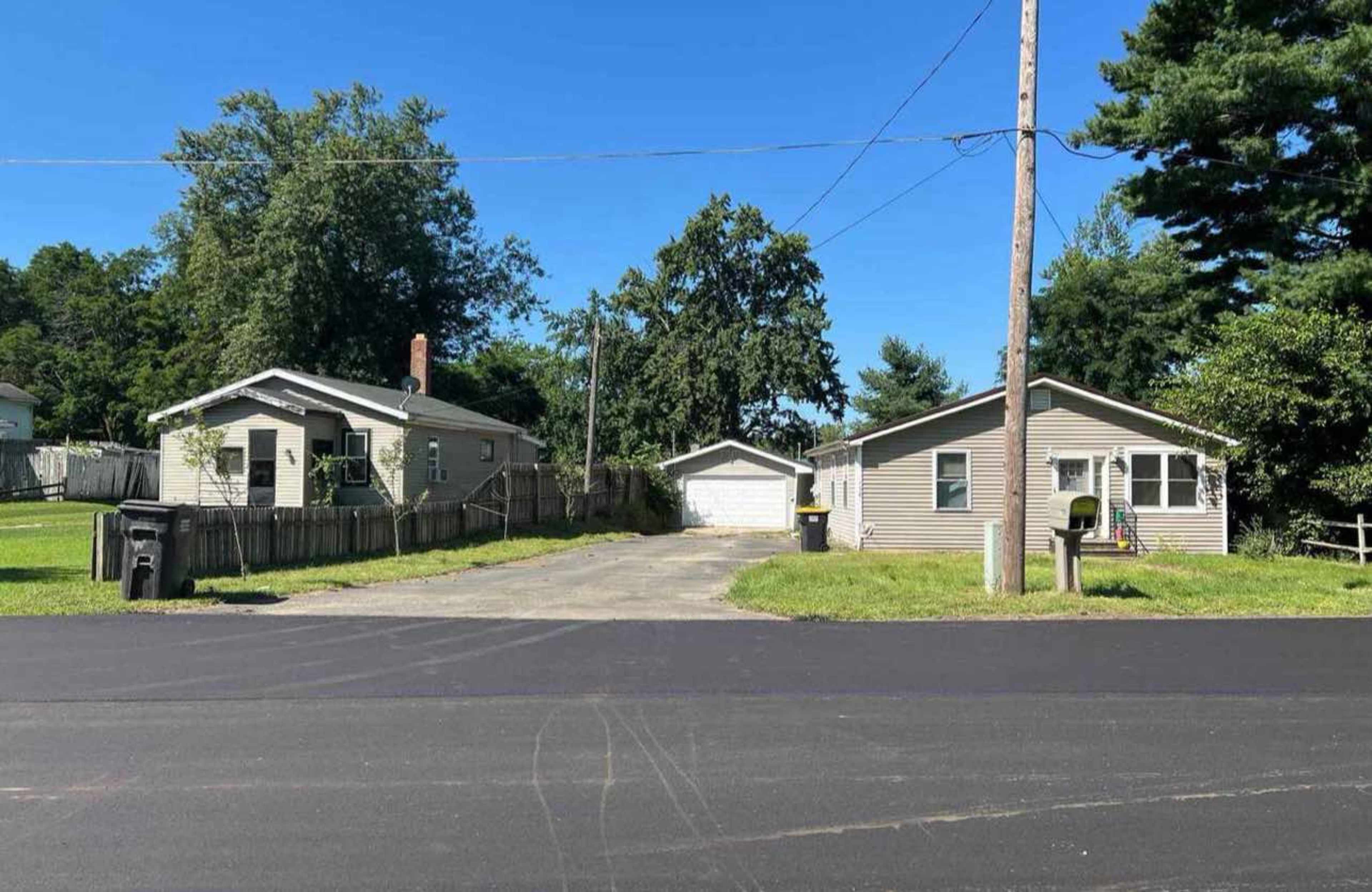 Two single-story houses are situated across from each other on a paved street, with a driveway leading to a garage and several trees in the background.
