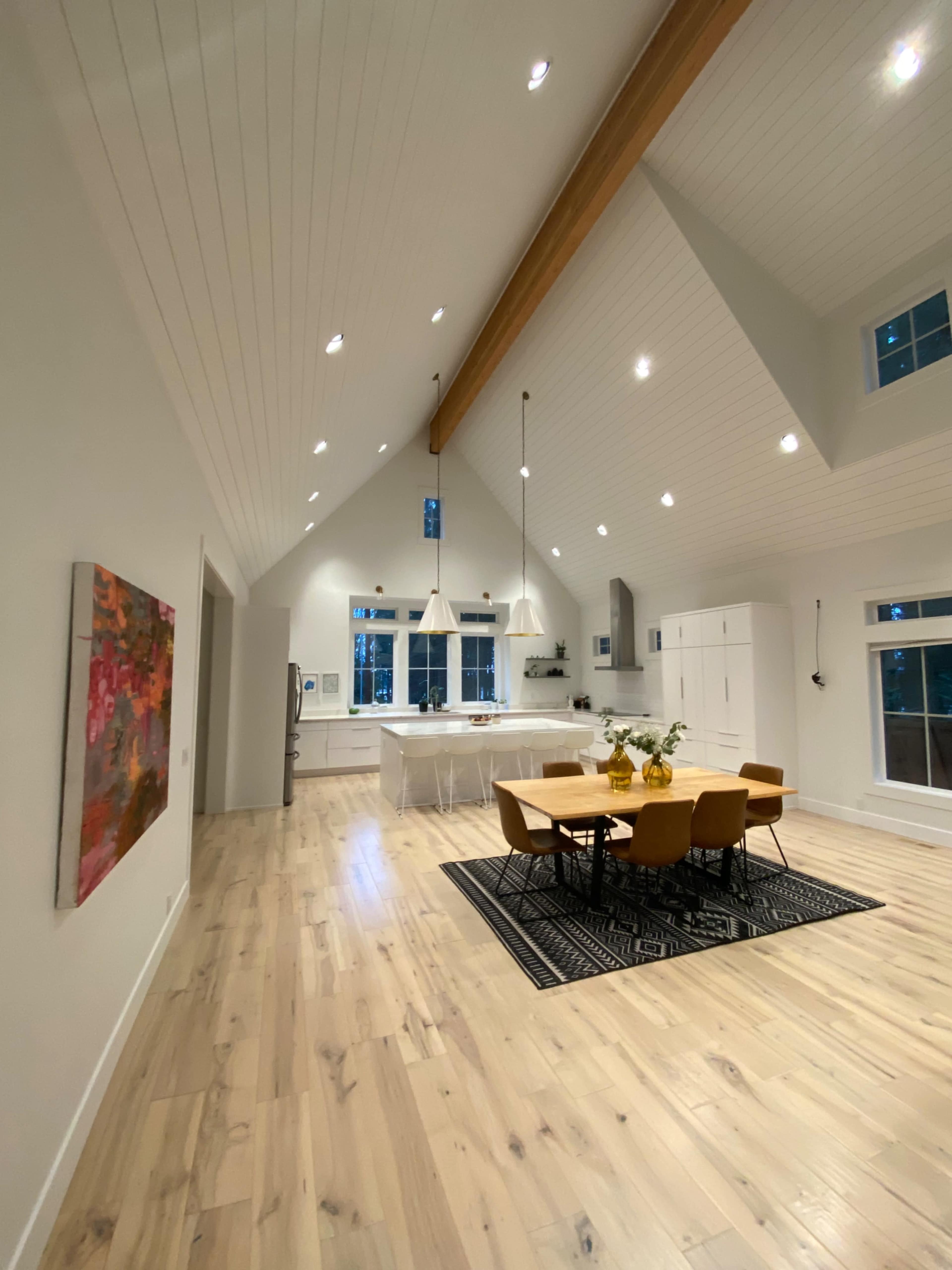 The image shows a spacious, well-lit kitchen and dining area with high ceilings, a wooden beam, and a modern dining table surrounded by yellow chairs.