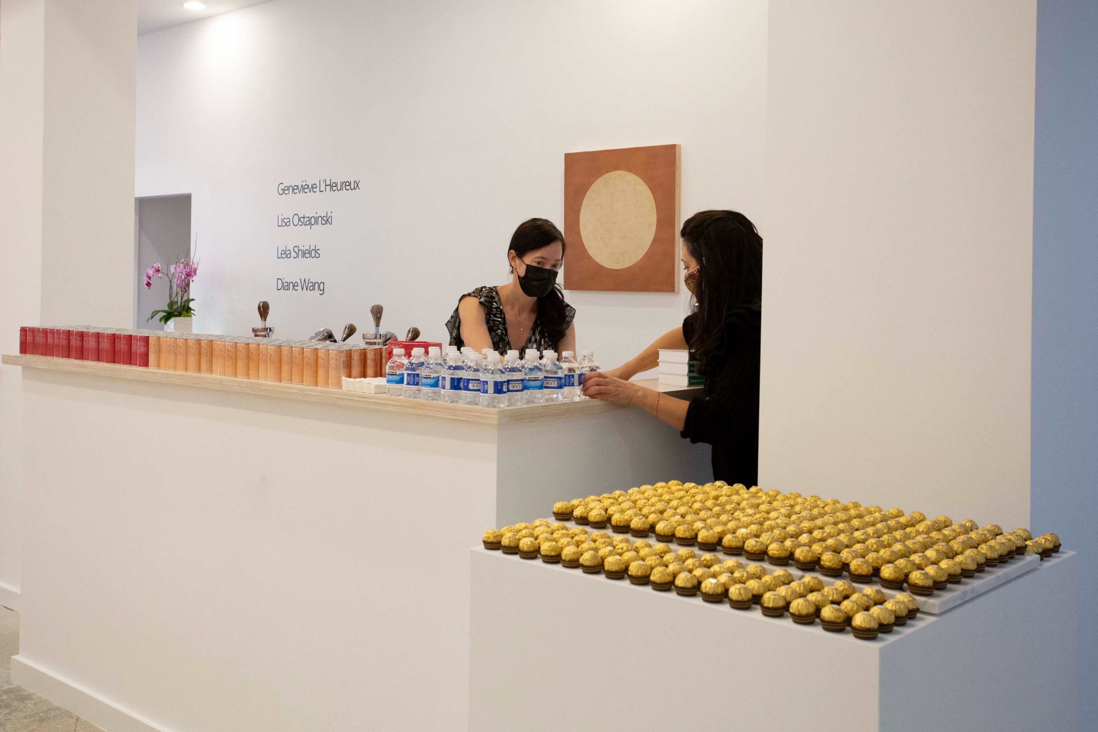 Two women in masks are arranging water bottles and a tray of golden desserts at a white reception counter, while a wall displays names and a round artwork.