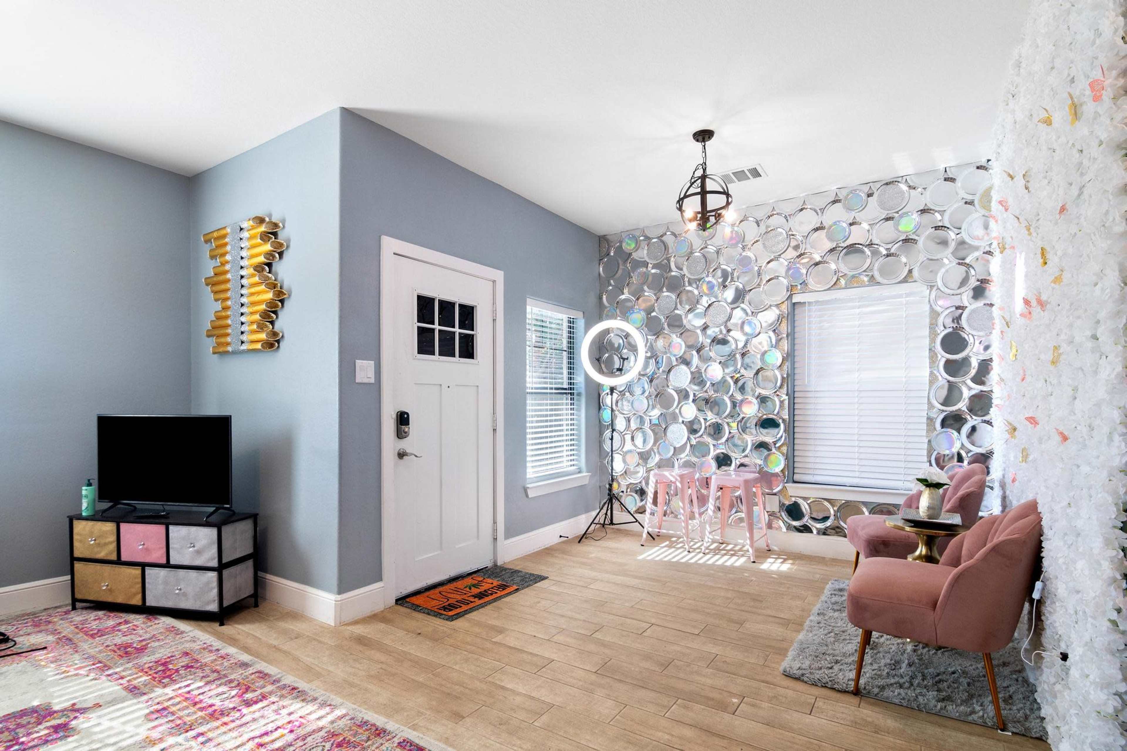The room features a light blue wall, a television on a stand, a round mirror backdrop, a small pink table with chairs, and a pair of pink armchairs.