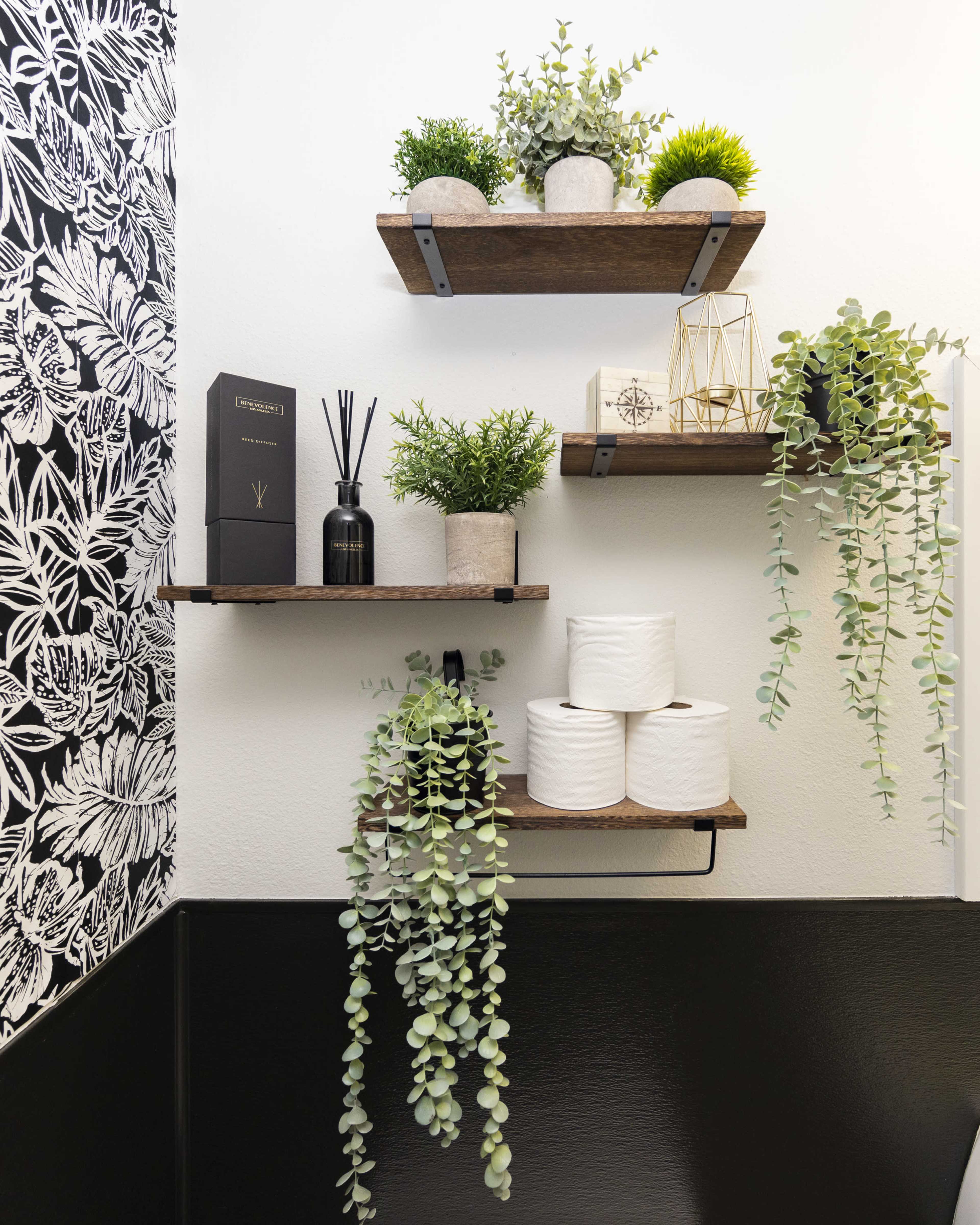 The image shows a corner with three wooden shelves displaying various plants and decorative items, alongside a holder with rolls of toilet paper.
