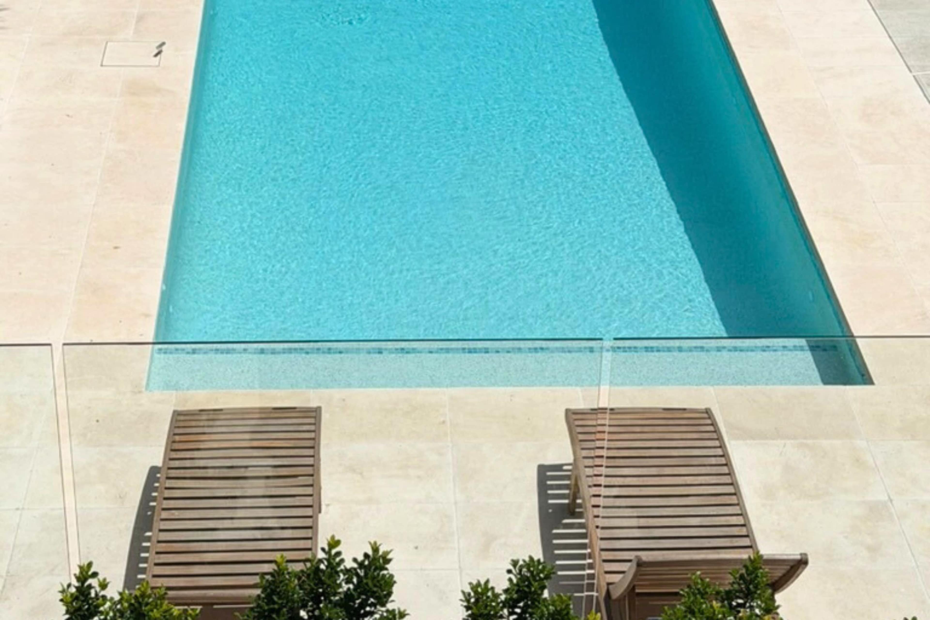 The image shows a rectangular swimming pool surrounded by a stone deck, with two wooden lounge chairs positioned on one side.