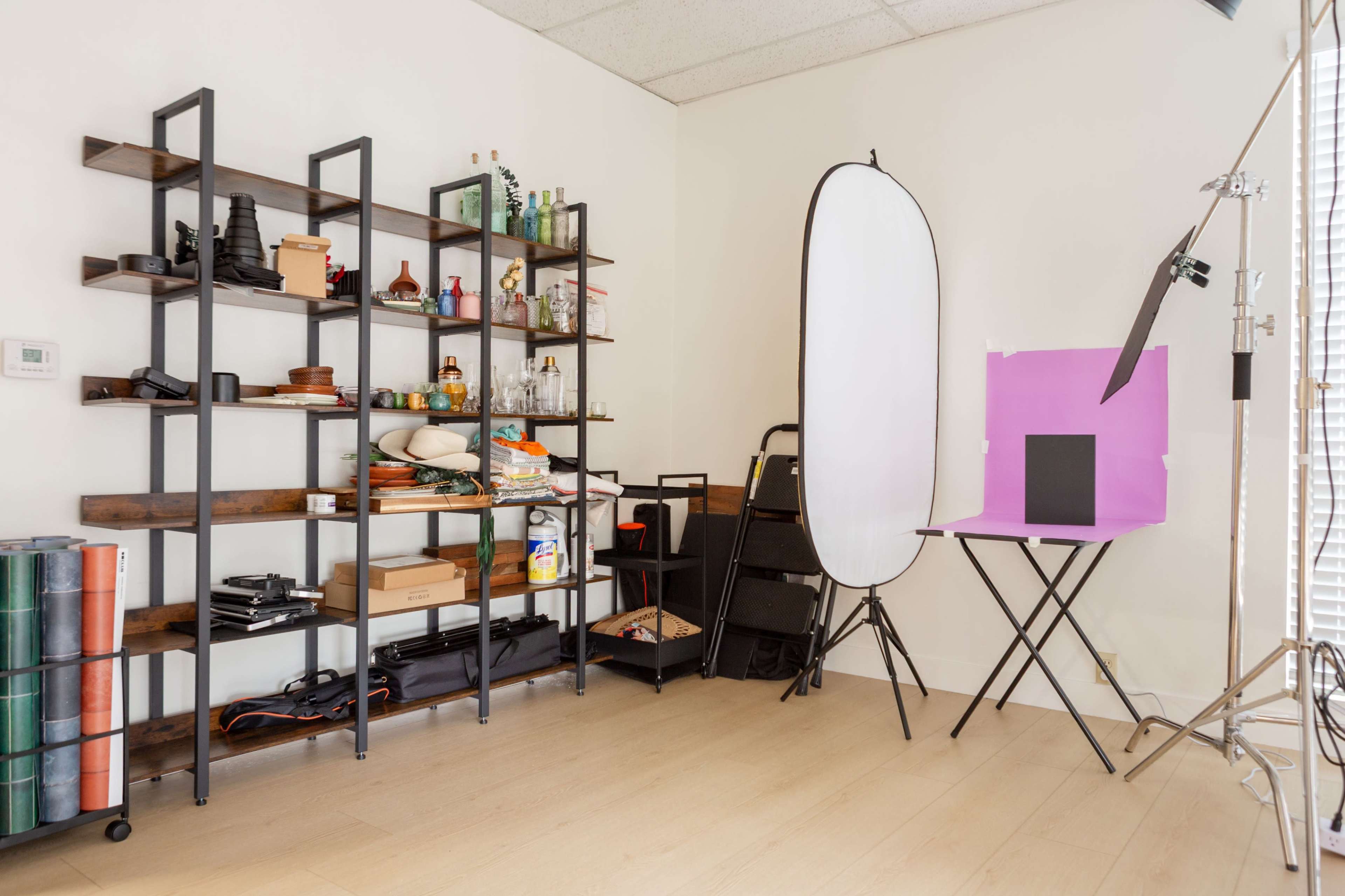 The image shows a photography studio setup featuring a shelving unit with various items, a backdrop stand with a white reflector, and a table holding a black backdrop.