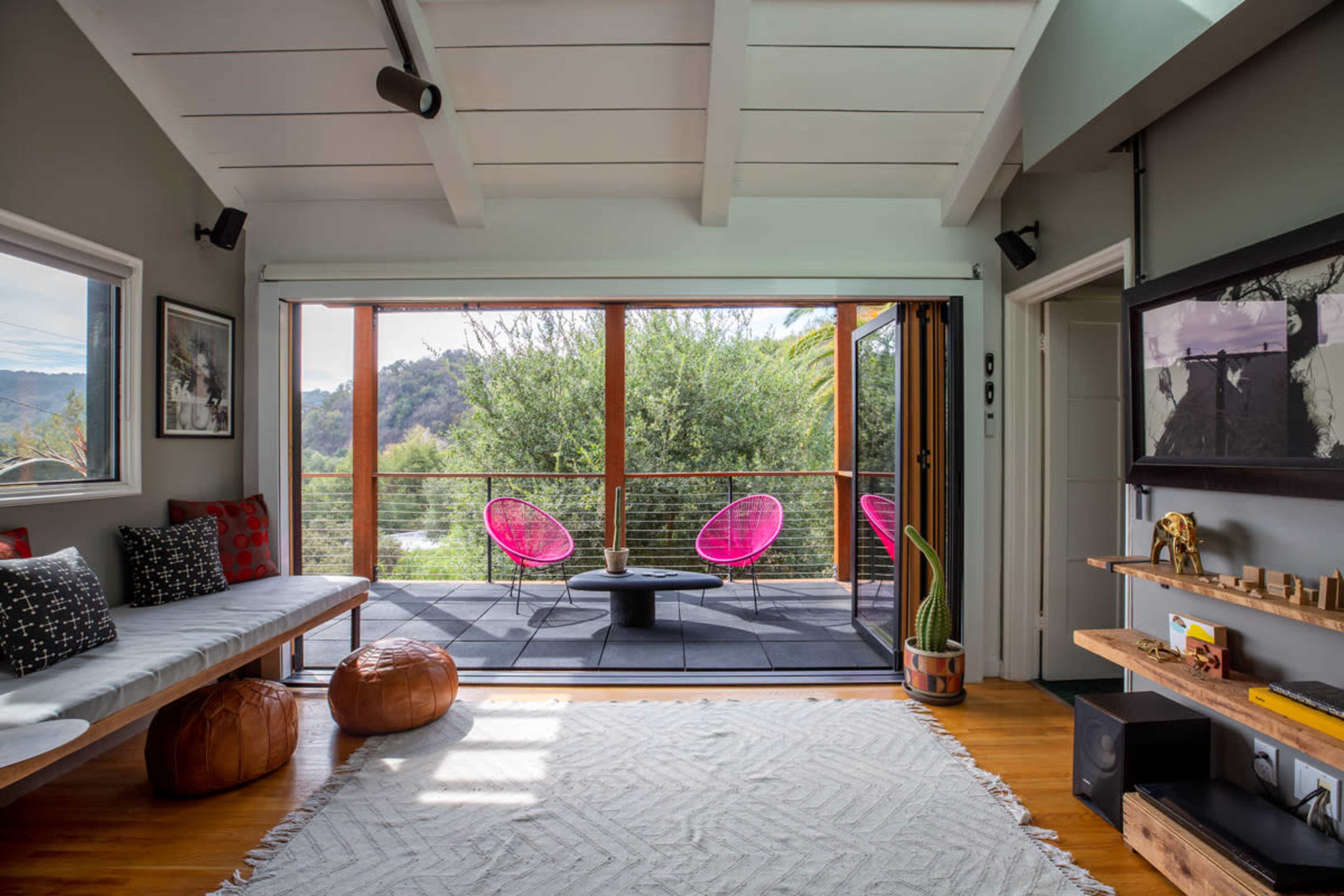 A modern living room with a large sliding door that opens to a deck featuring two pink chairs and a view of greenery outside.