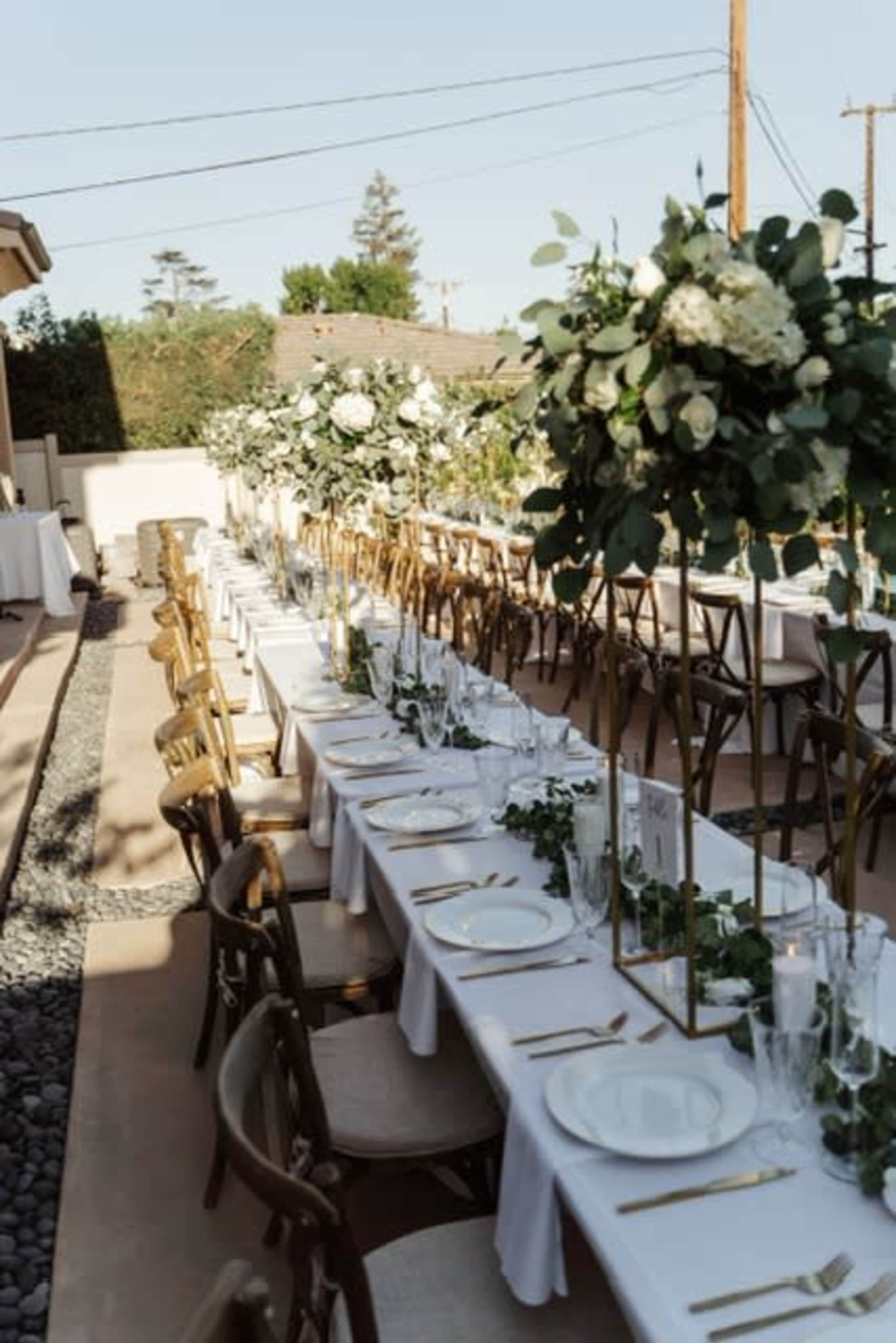A long, elegantly set outdoor dining table features tall floral centerpieces and is surrounded by wooden chairs.