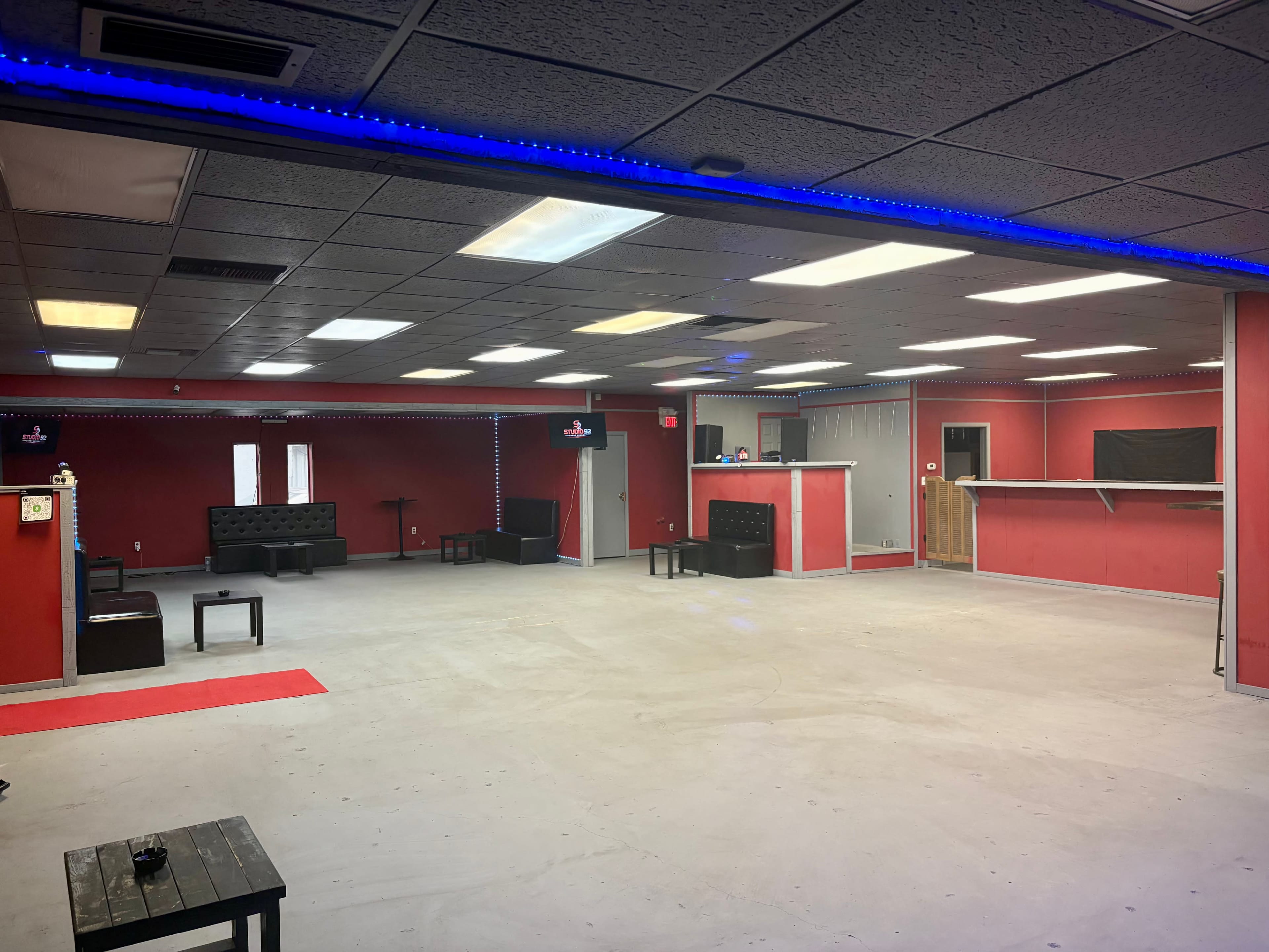 The image shows an empty interior space with red walls, a few black seating areas, and a bar counter in the background.