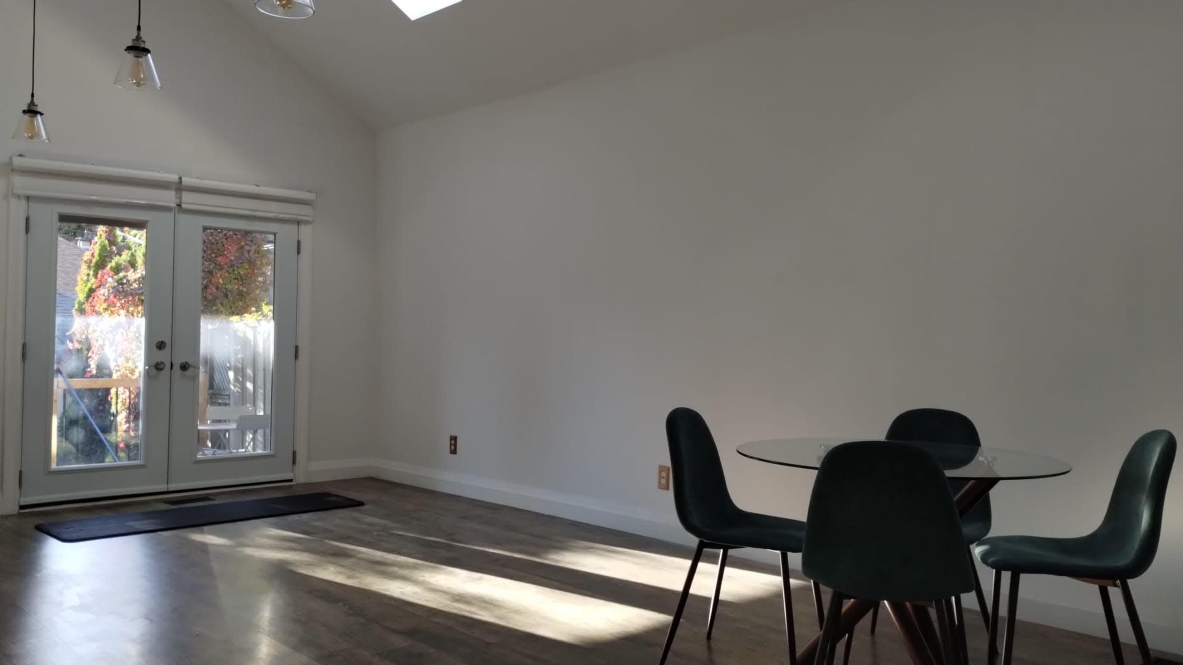 The image shows a minimalist dining area with a glass table and four green chairs, adjacent to a set of glass doors leading to an outdoor space.