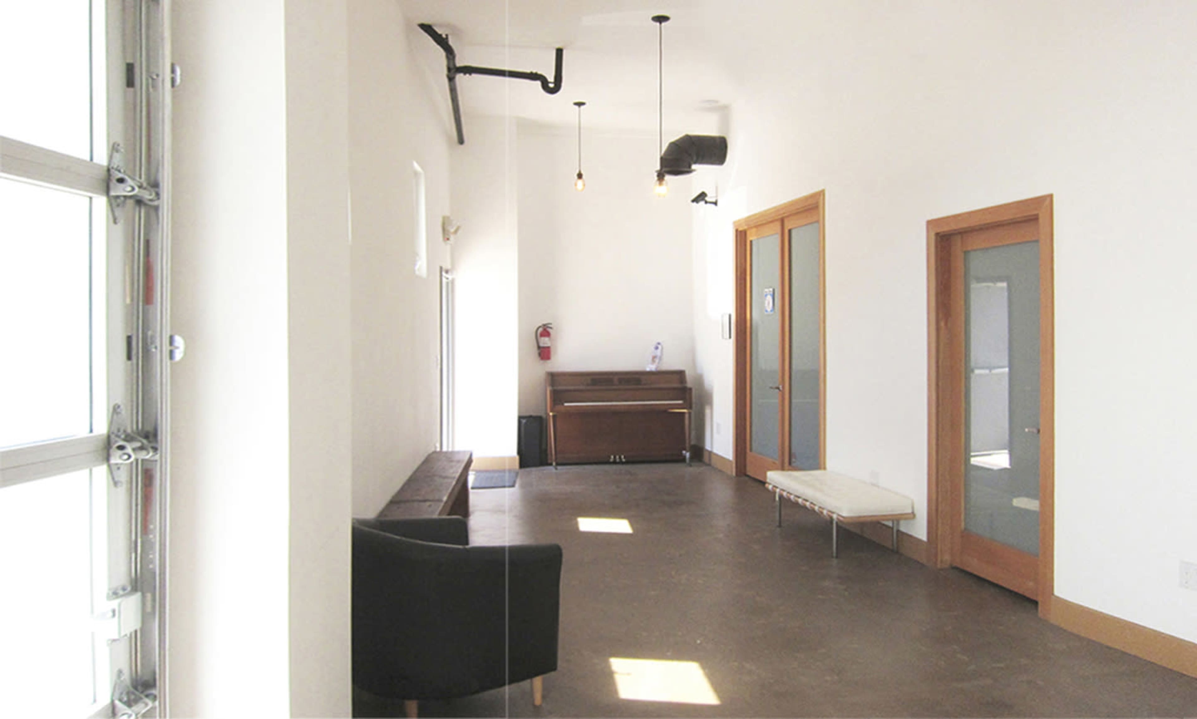 The image depicts a spacious, minimalist interior with polished concrete flooring, a piano in the corner, and wooden accents on the doors and furniture.