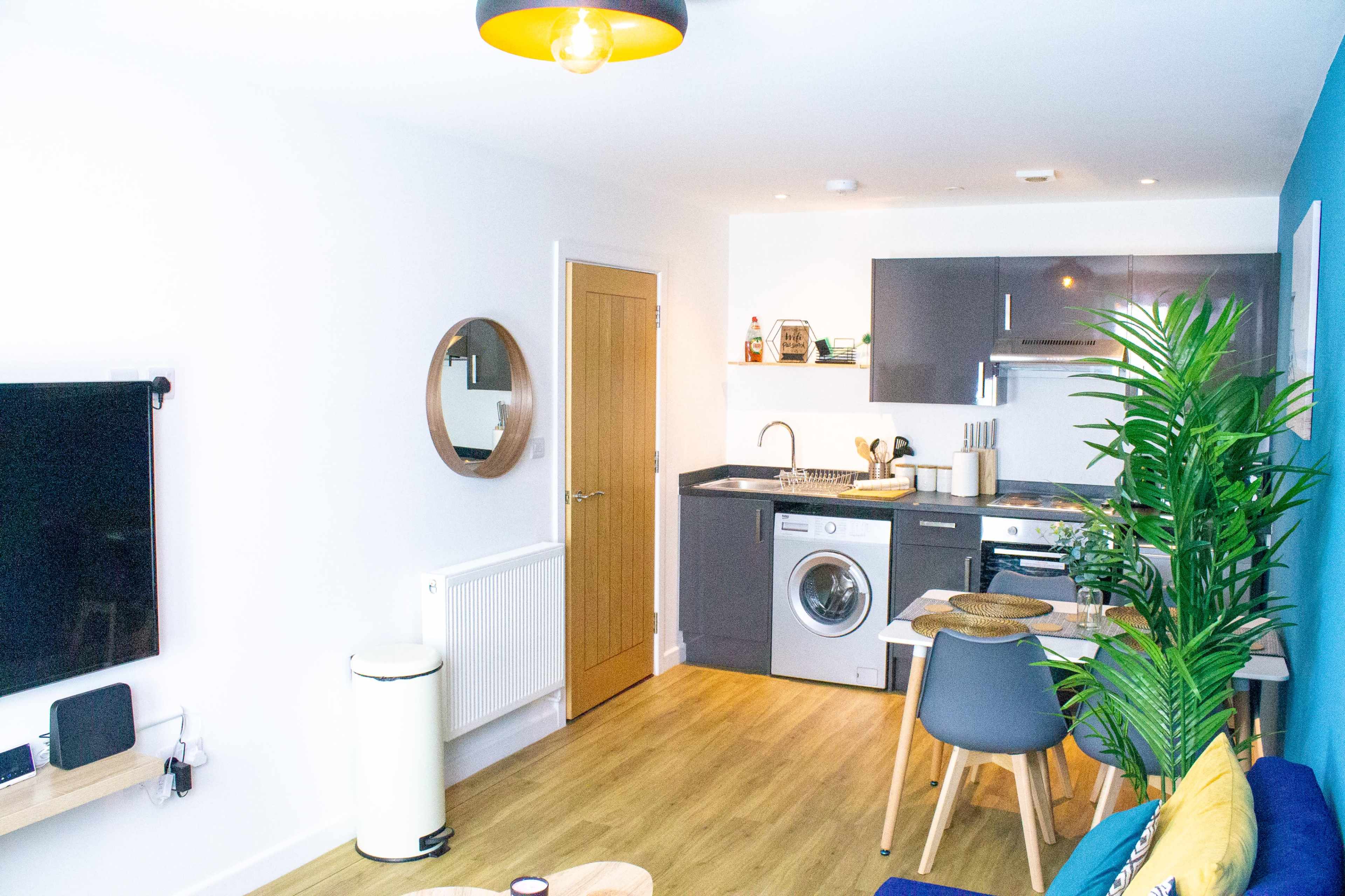 A modern, open-plan living space featuring a small kitchenette, a washing machine, and a seating area with a blue sofa and a round coffee table.