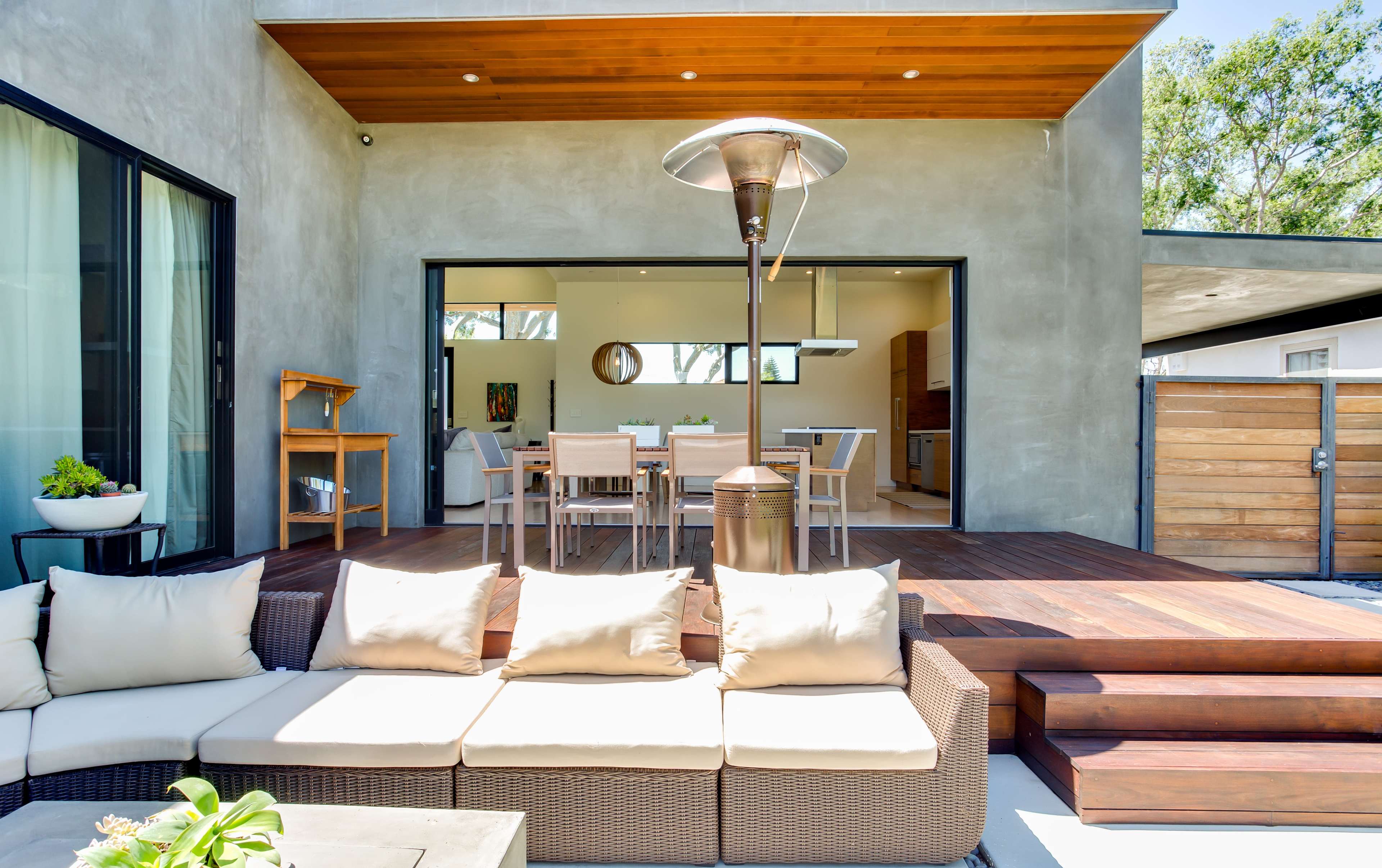 A modern outdoor seating area features a sectional sofa and a patio heater, overlooking an open-plan dining space through large sliding doors.