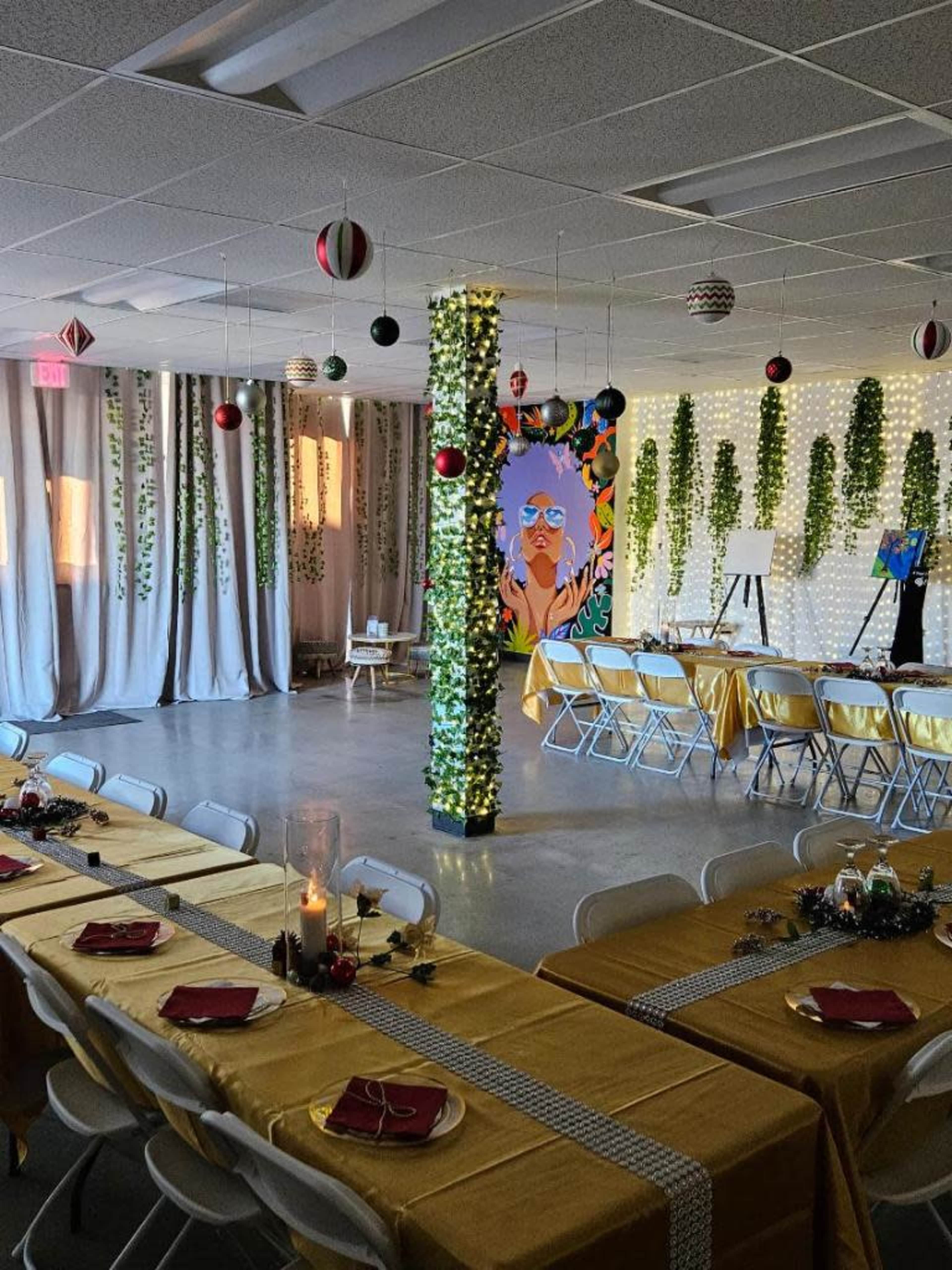A decorated indoor event space featuring rows of tables with yellow tablecloths, hanging ornaments, and greenery on the walls and pillars.