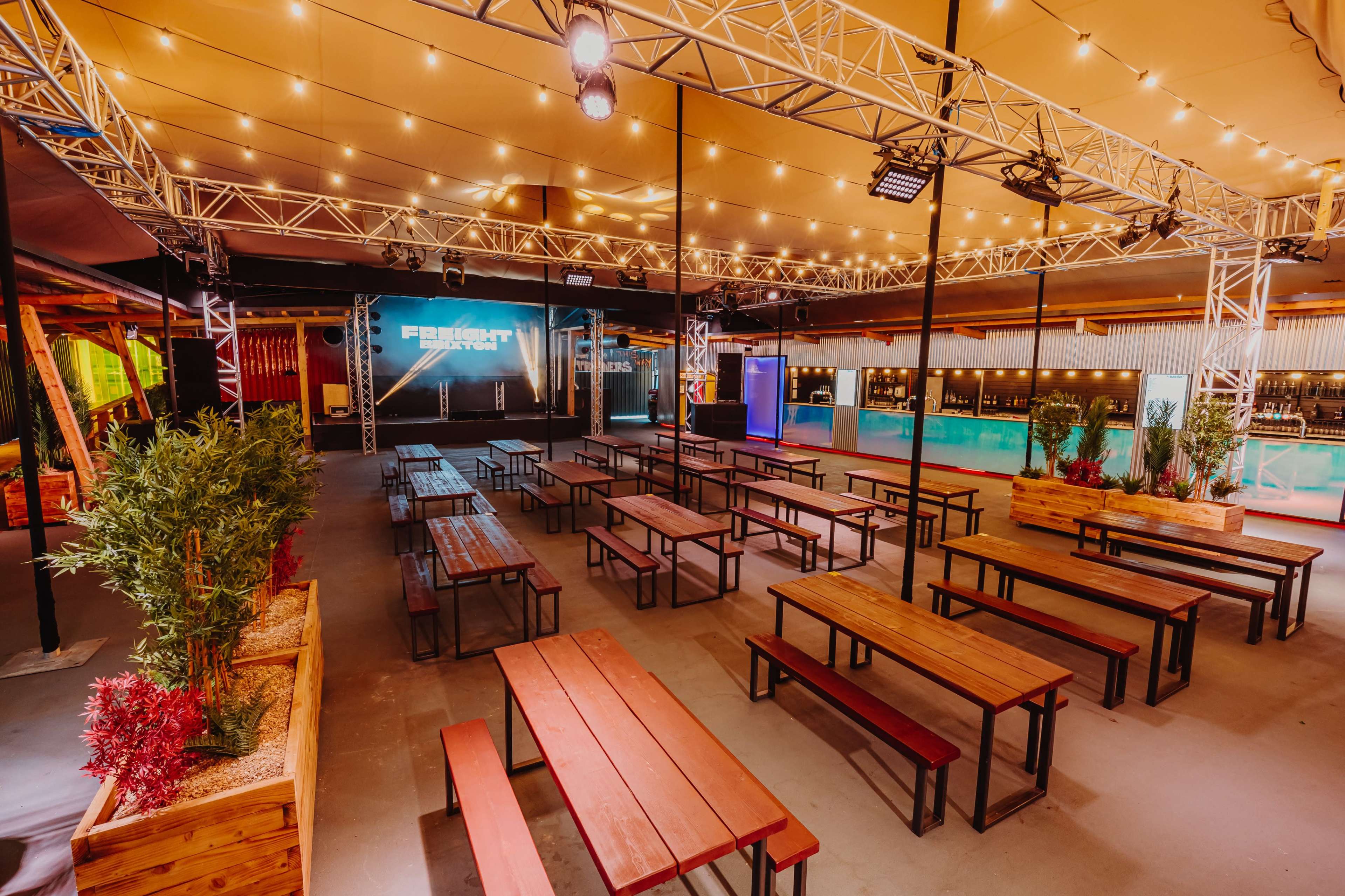 The image shows an indoor venue with a stage and rows of wooden benches arranged around a bar area, all under warm lighting.