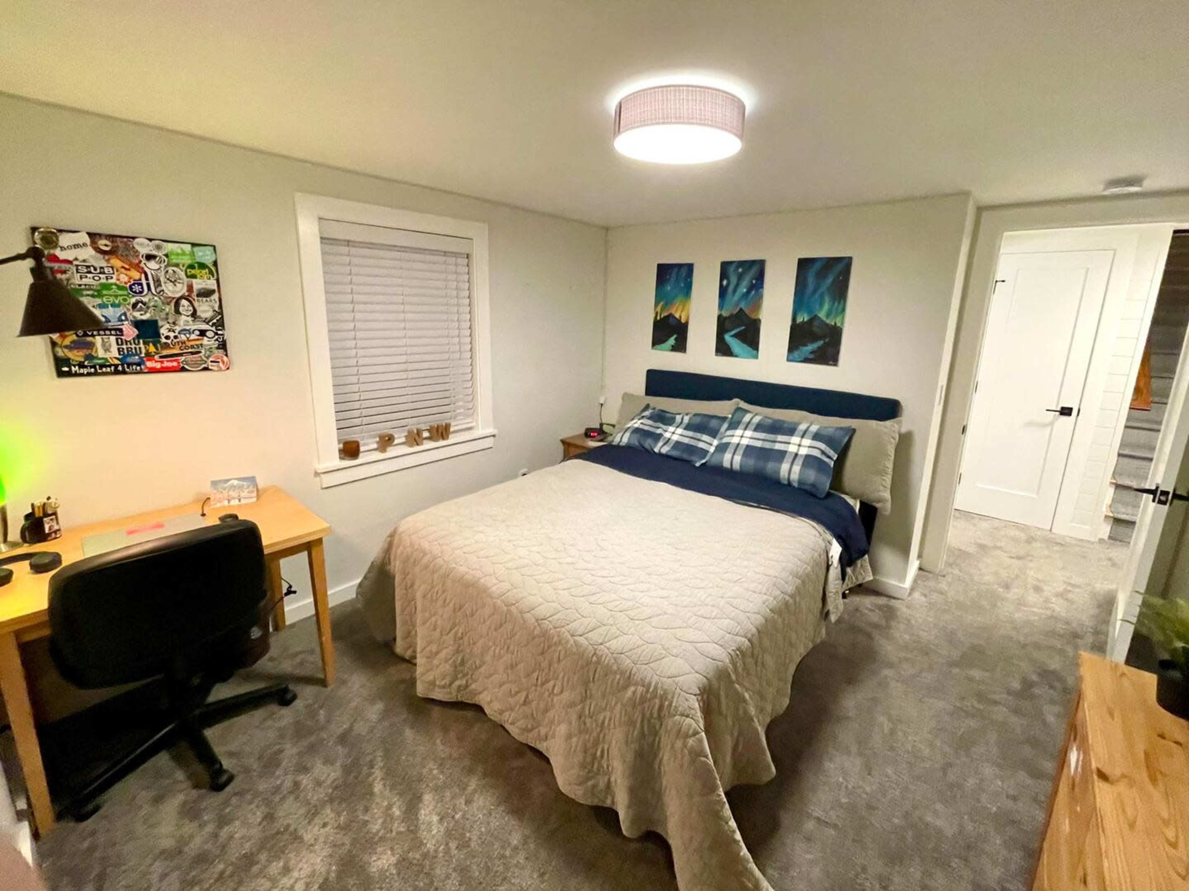 A well-lit bedroom featuring a large bed with a quilted cover, a desk with a chair, and three framed pictures on the wall.